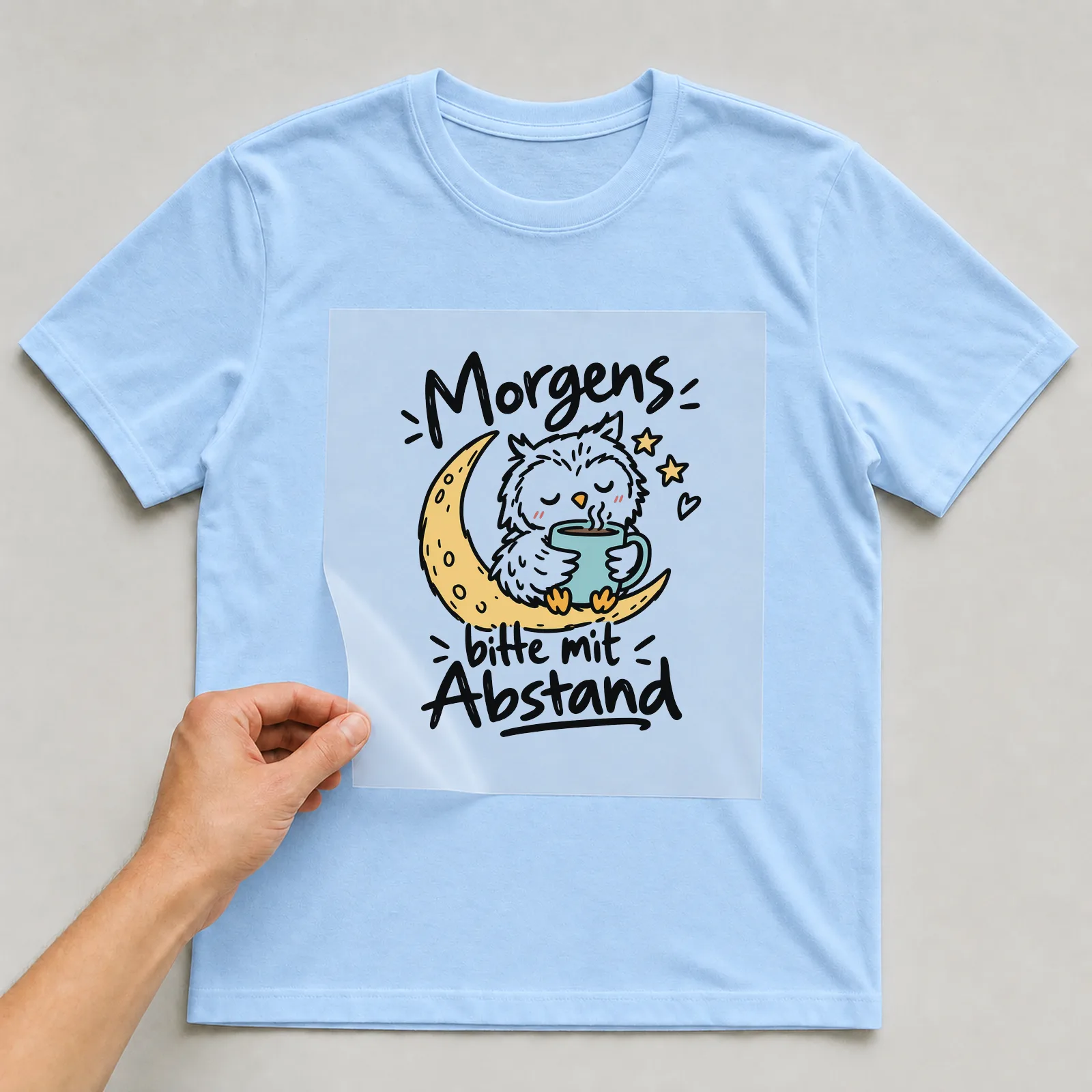 Bügelbild „Morgens bitte mit Abstand“ auf hellblauem T-Shirt, Folie wird aufgelegt
