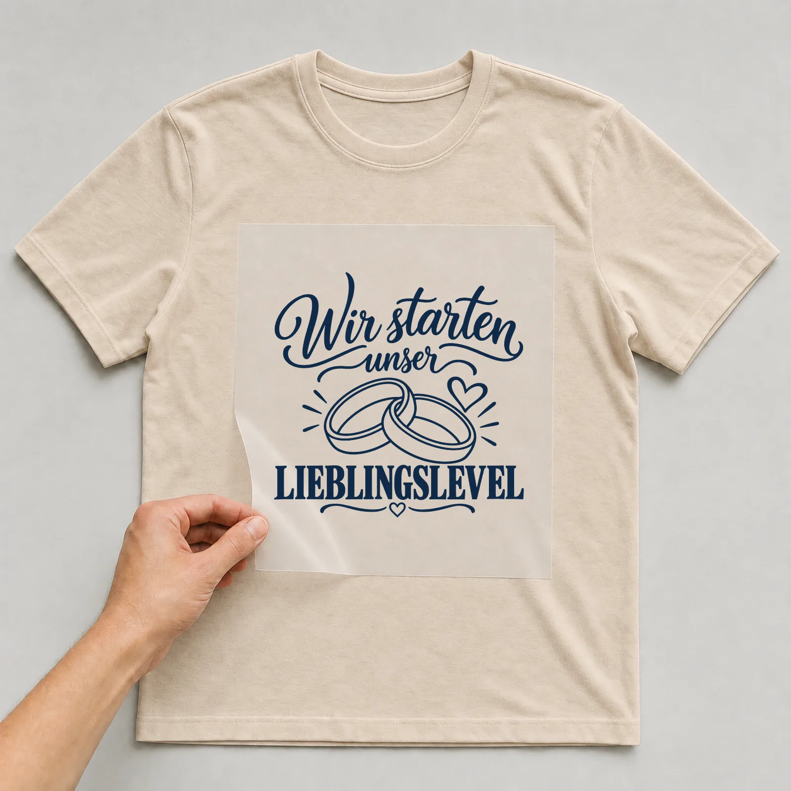 Bügelbild „Wir starten unser Lieblingslevel“ auf beigem T-Shirt, Folie wird aufgelegt