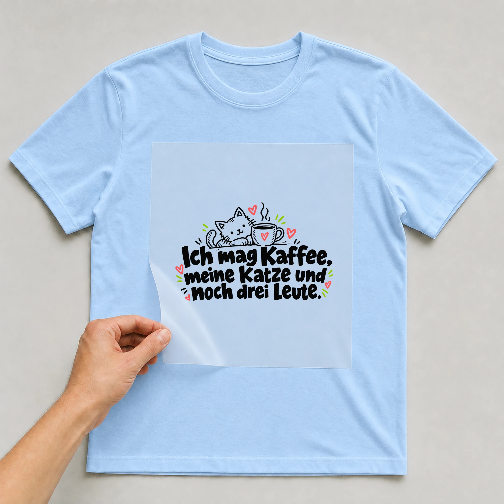 Bügelbild „Ich mag Kaffee, meine Katze und noch drei Leute.“ auf babyblauem T-Shirt, Folie wird aufgelegt
