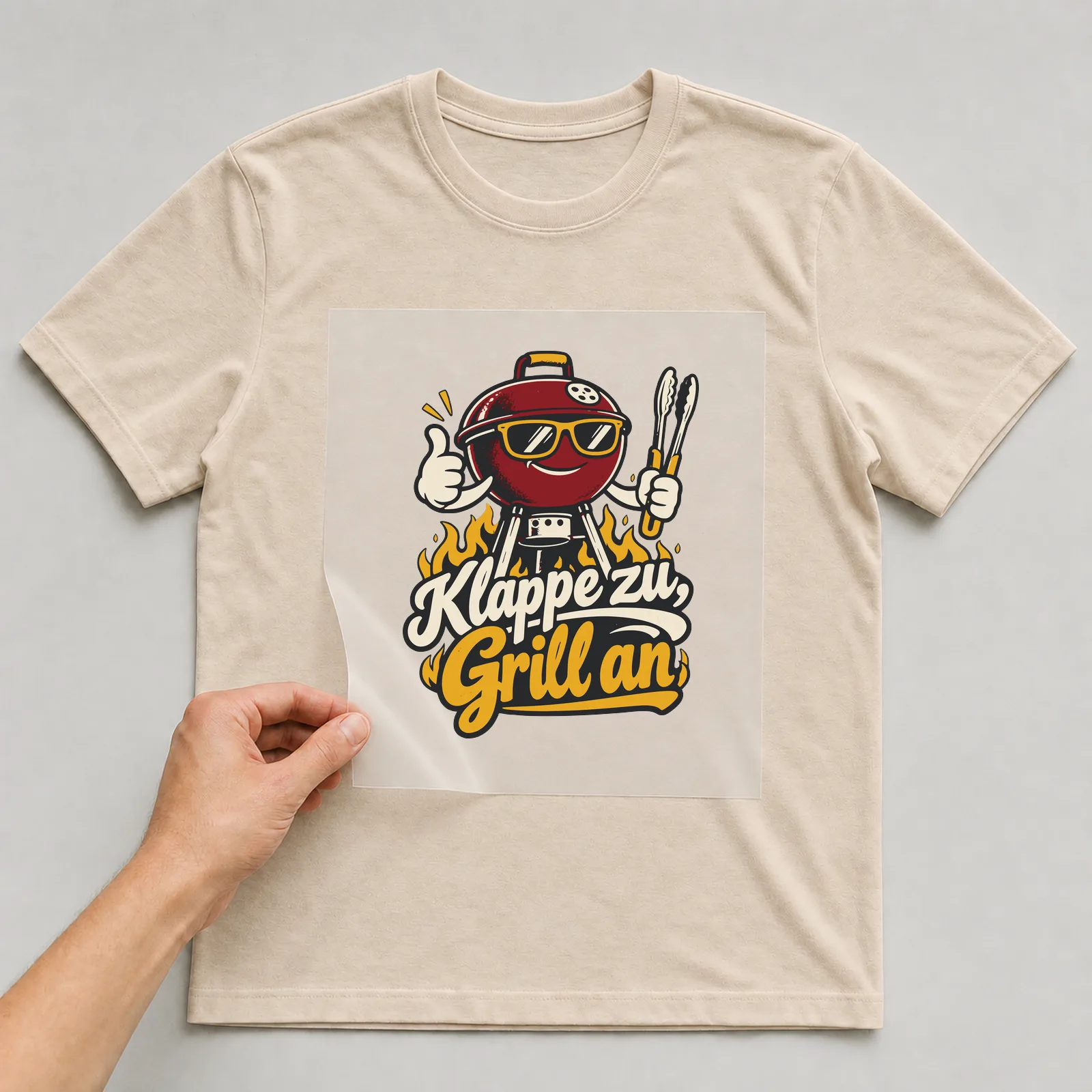 Bügelbild Klappe zu, Grill an auf beigem T-Shirt, Folie wird aufgelegt