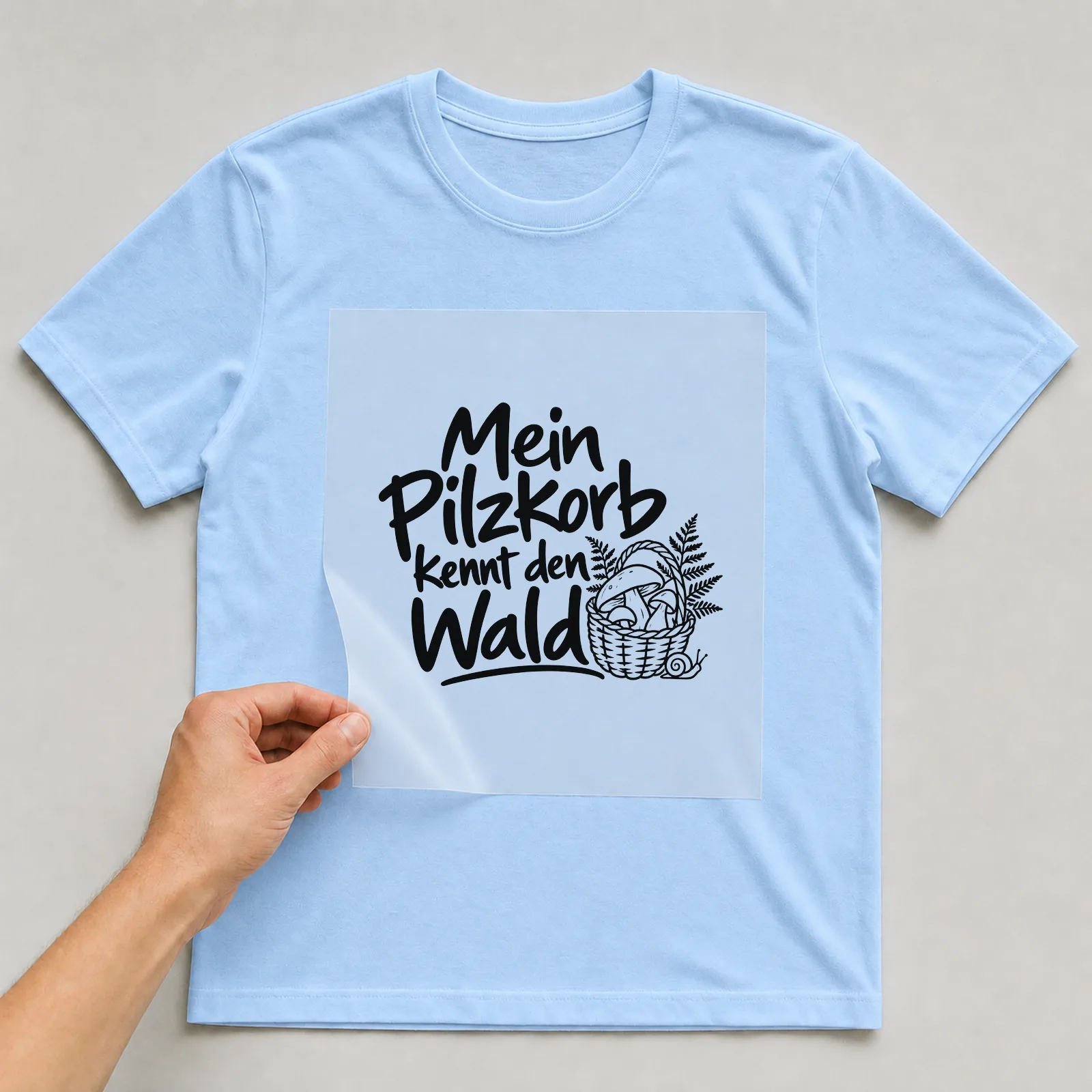 Bügelbild „Mein Pilzkorb kennt den Wald“ auf hellblauem T-Shirt, Folie wird aufgelegt