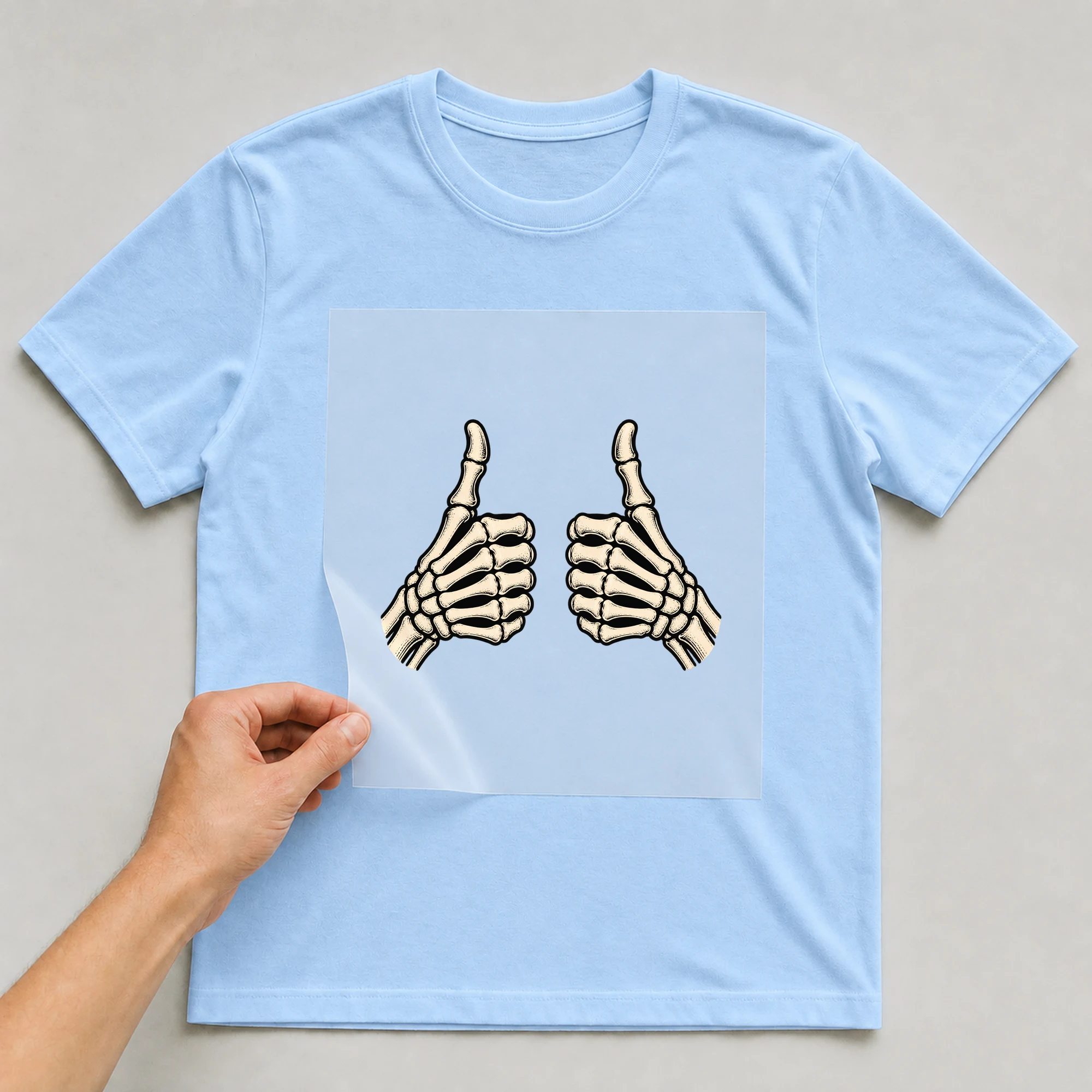 Bügelbild „Skelett-Hände mit Daumen hoch“ auf babyblauem T-Shirt, Folie wird aufgelegt