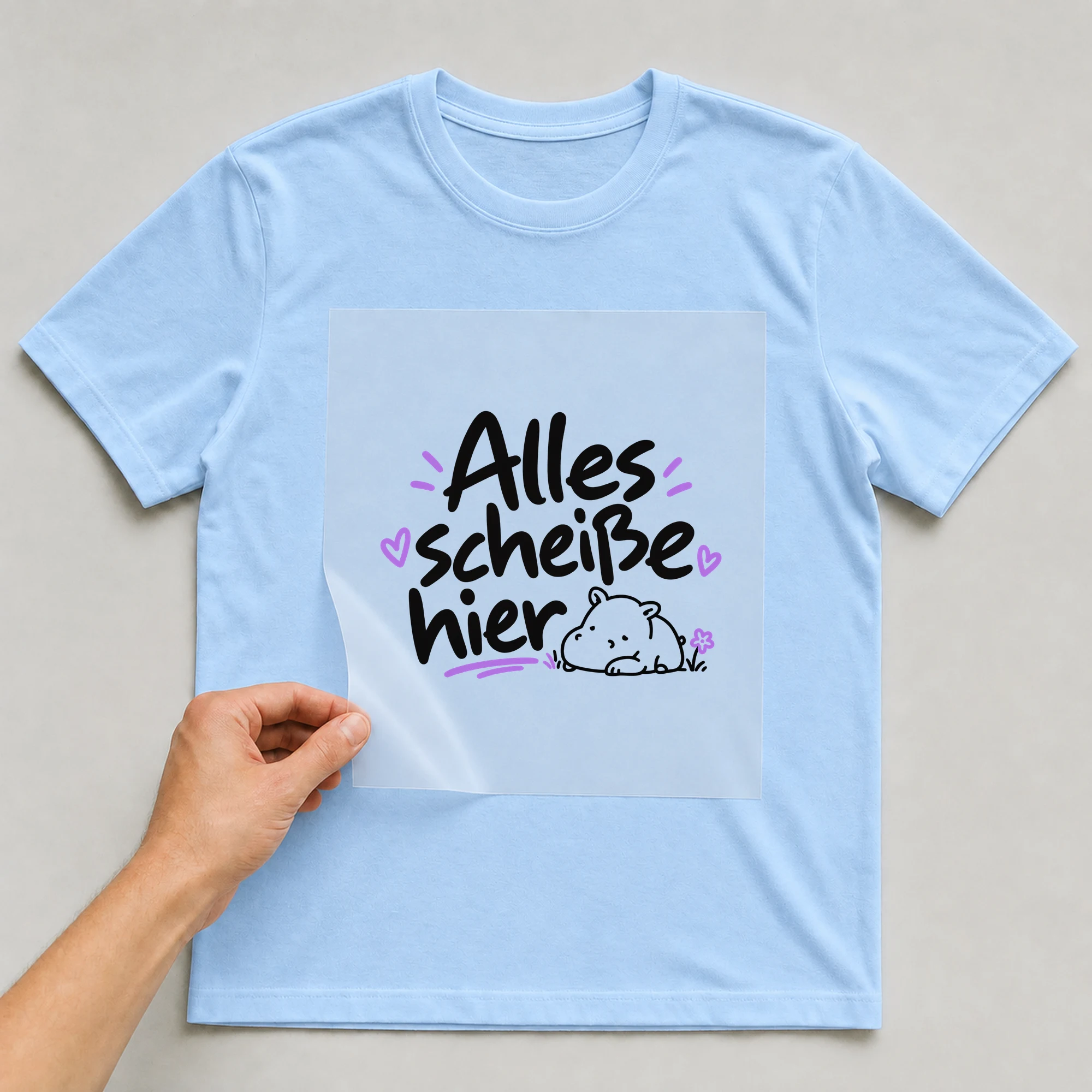 Bügelbild „Alles sch*iße hier“ auf babyblauem T-Shirt, Folie wird aufgelegt