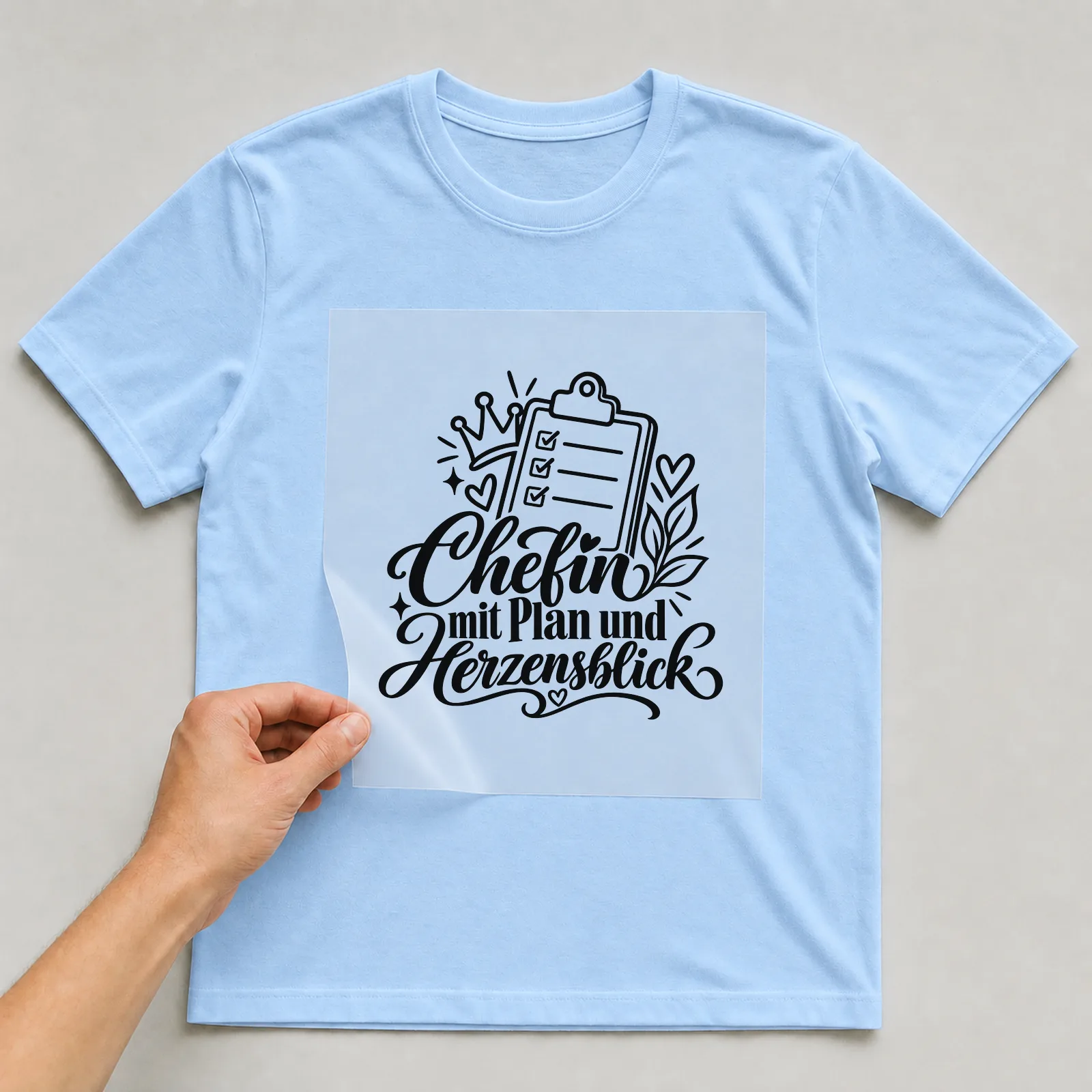 Bügelbild „Chefin mit Plan und Herzensblick“ auf hellblauem T-Shirt, Folie wird aufgelegt