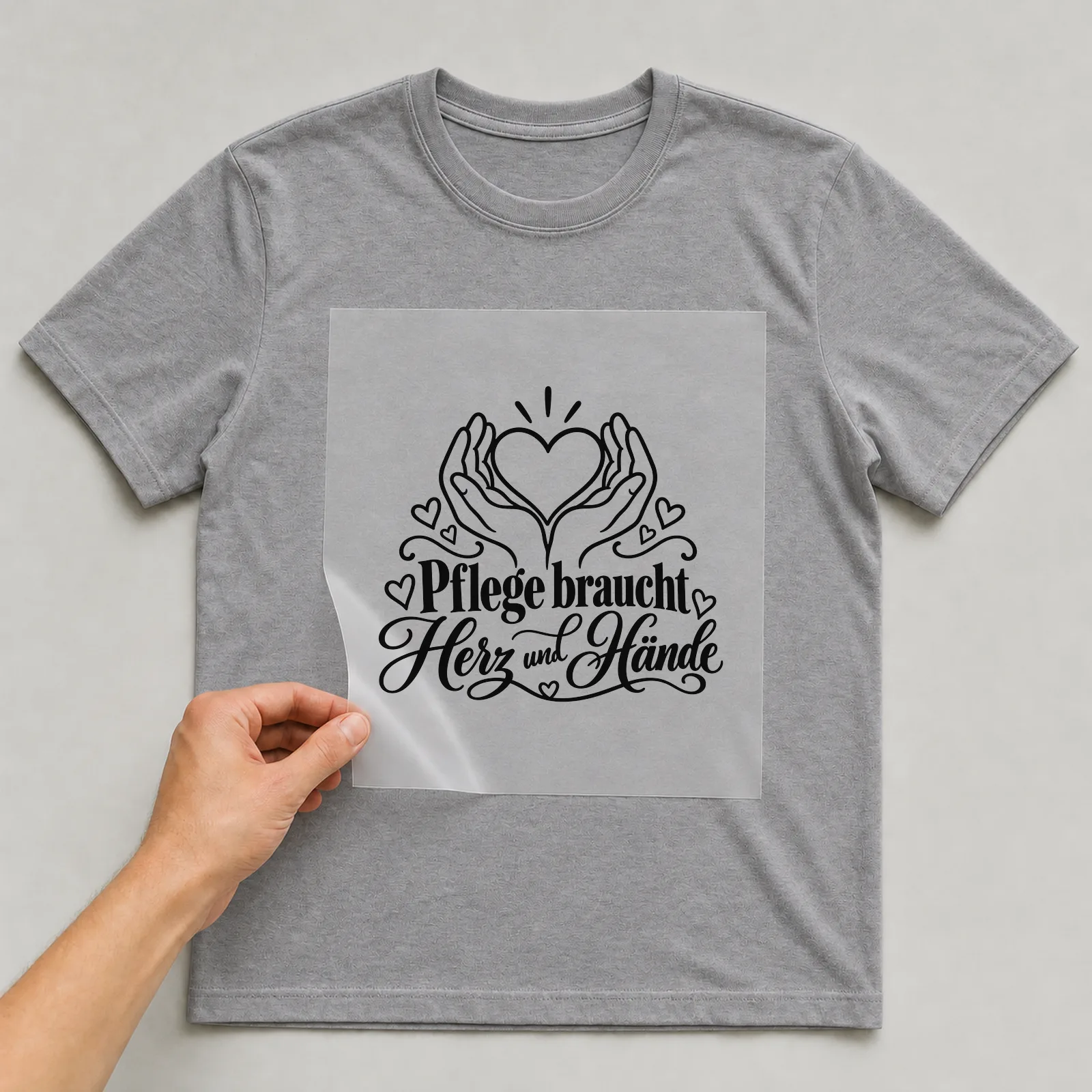 Bügelbild „Pflege braucht Herz und Hände“ auf grauem T-Shirt, Folie wird aufgelegt