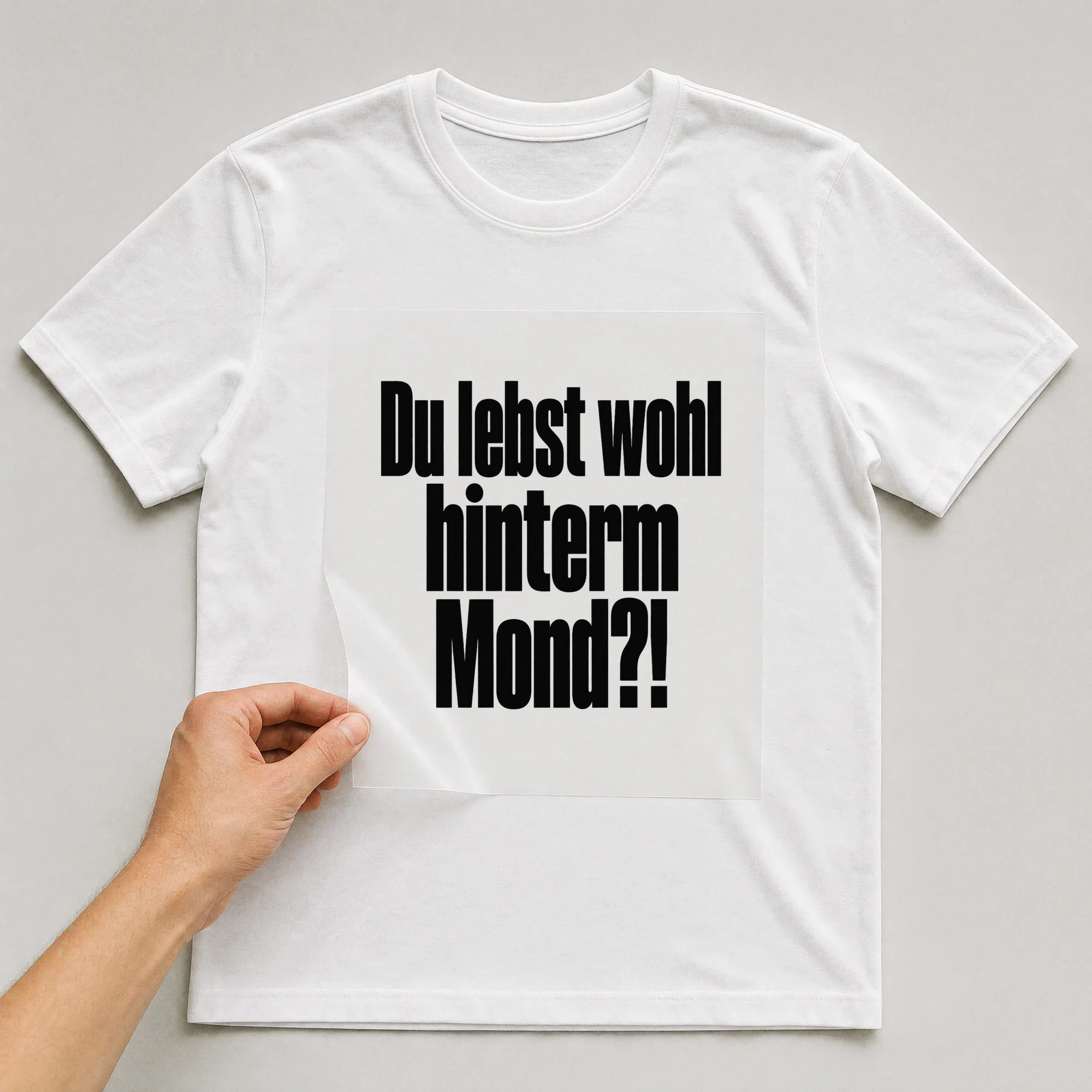 Bügelbild „Du lebst wohl hinterm Mond?!“ auf weißem T-Shirt, Folie wird aufgelegt