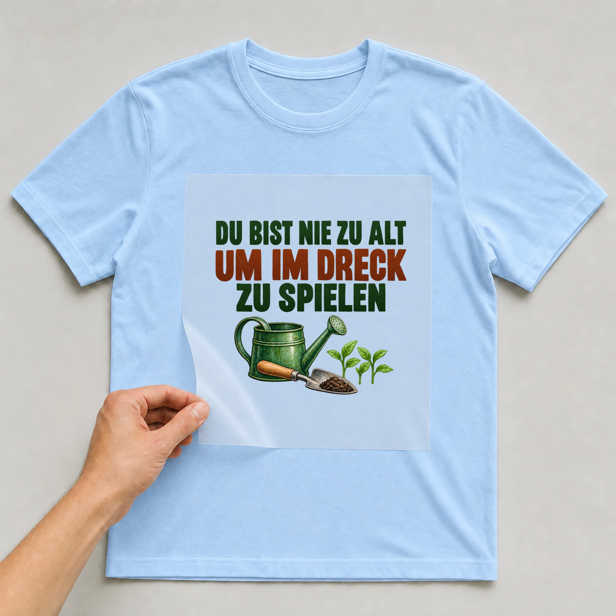 Bügelbild „Du bist nie zu alt um im Dreck zu spielen“ auf babyblauem T-Shirt, Folie wird aufgelegt