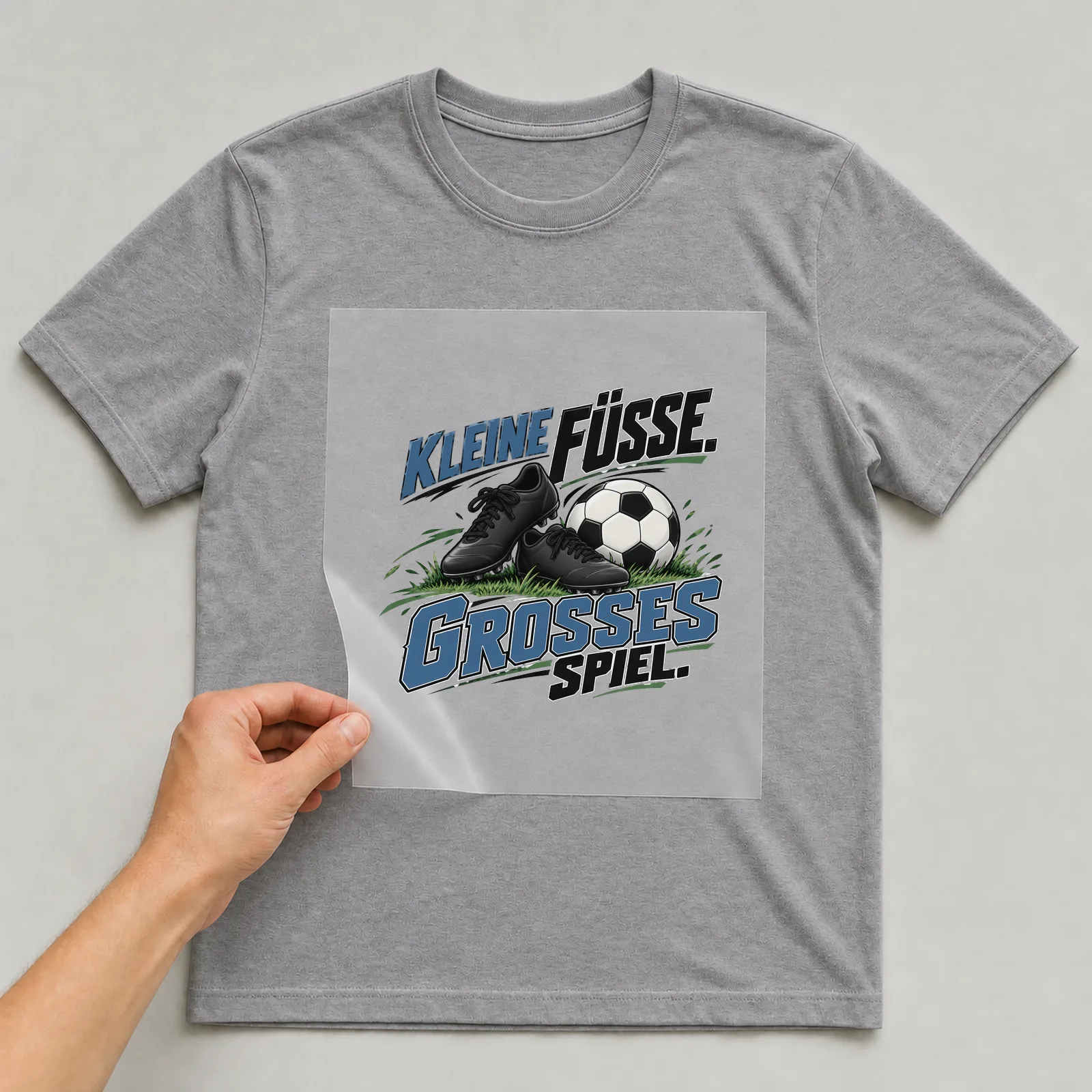 Bügelbild „Kleine Füße. Großes spiel.“ auf grauem T-Shirt, Folie wird aufgelegt
