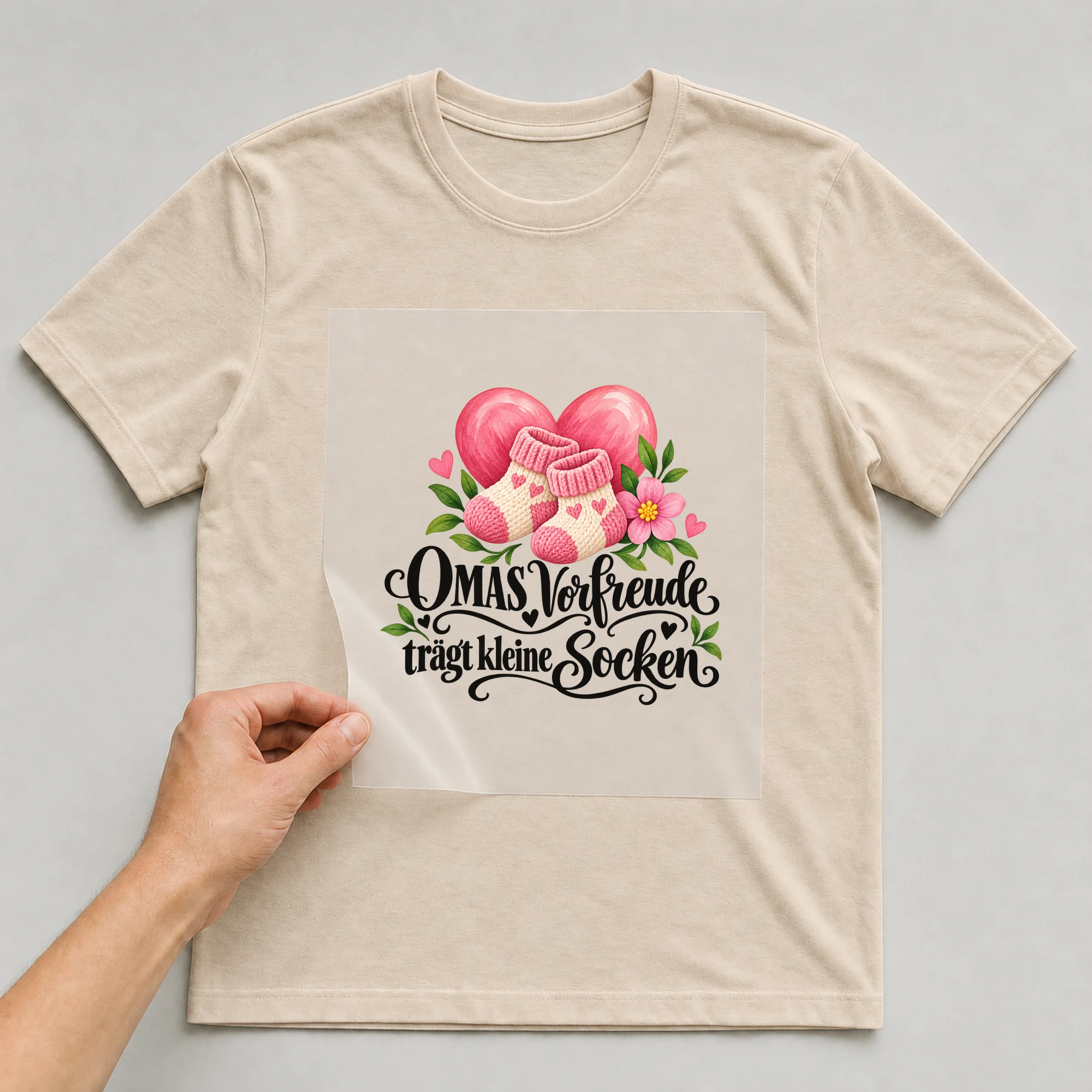 Bügelbild „OMAS Vorfreude trägt kleine Socken“ auf beigem T-Shirt, Folie wird aufgelegt