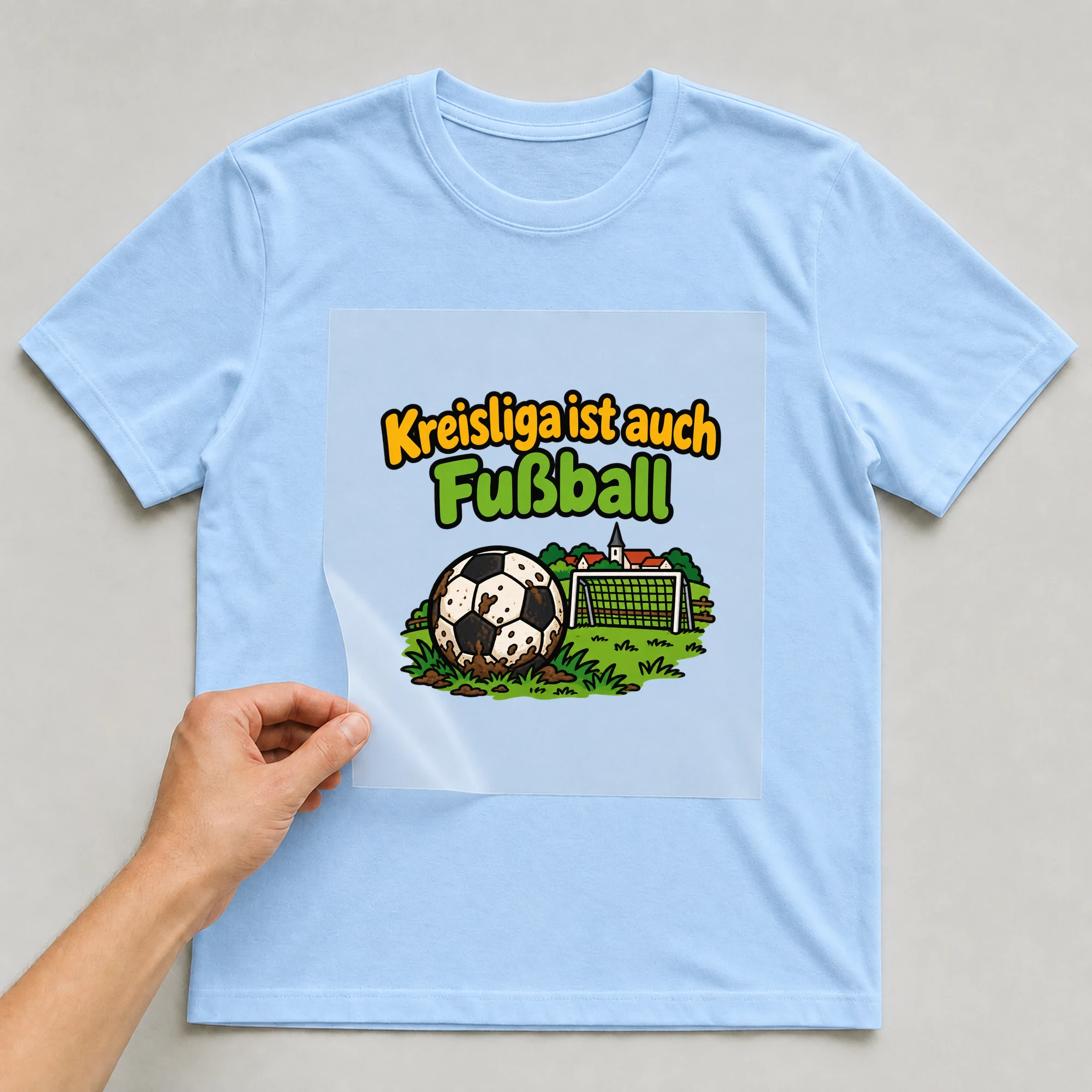 Bügelbild „Kreisliga ist auch Fußball“ auf babyblauem T-Shirt, Folie wird aufgelegt