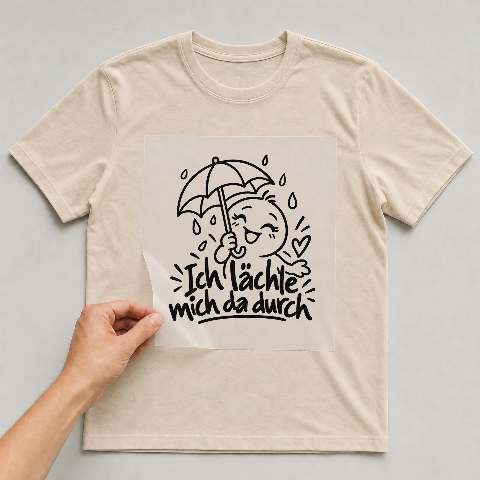 Bügelbild „Ich lächle mich da durch“ auf beigem T-Shirt, Folie wird aufgelegt