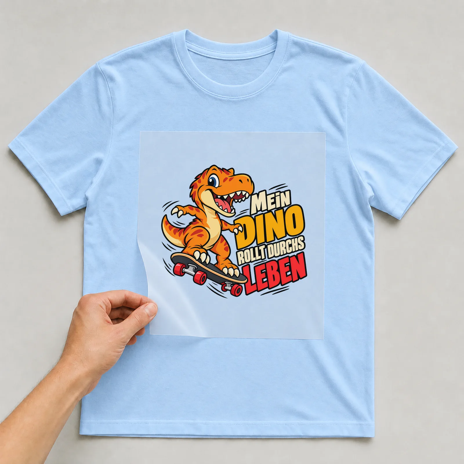 Bügelbild „Mein Dino rollt durchs Leben“ auf hellblauem T-Shirt, Folie wird aufgelegt