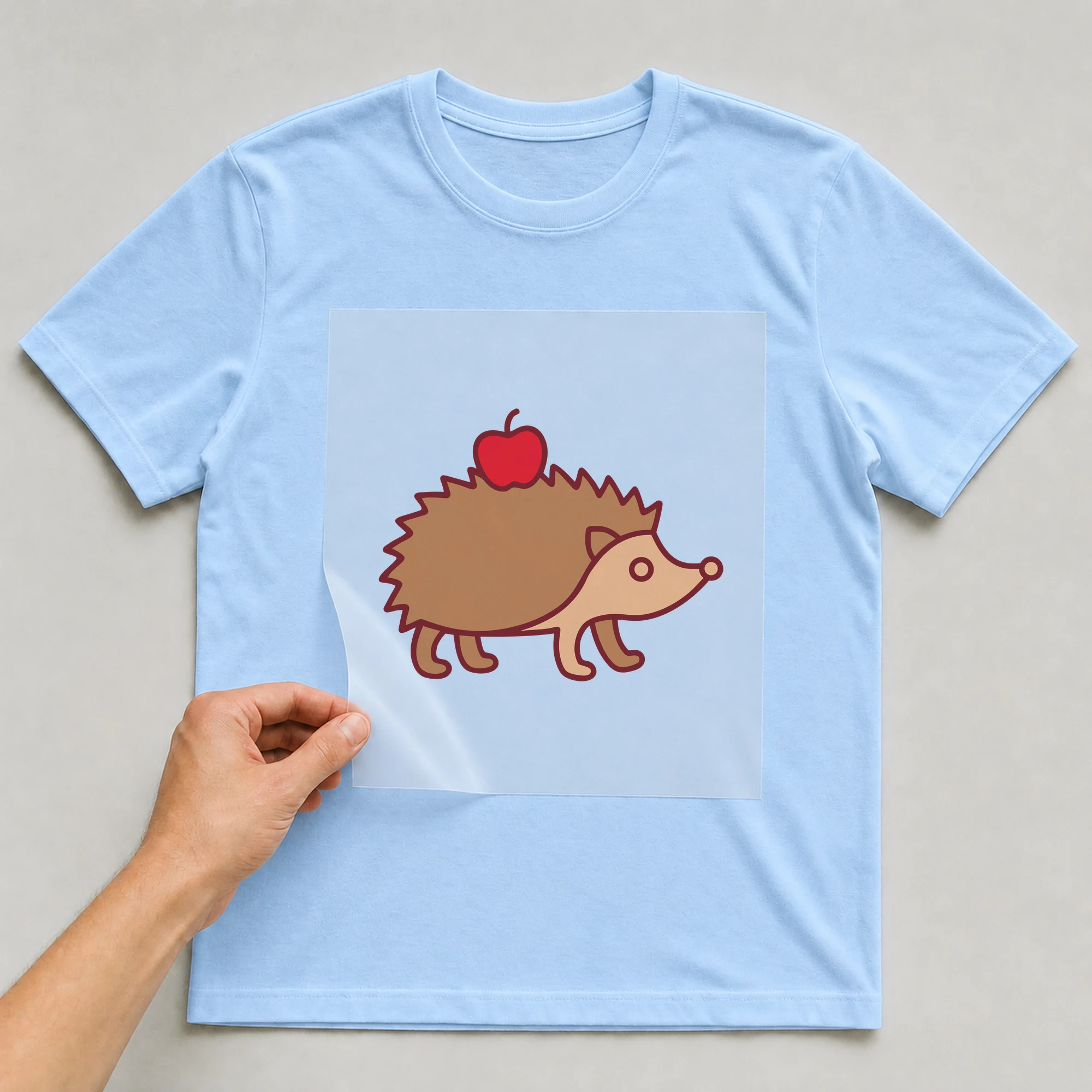Bügelbild „Igel mit Apfel“ auf babyblauem T-Shirt, Folie wird aufgelegt