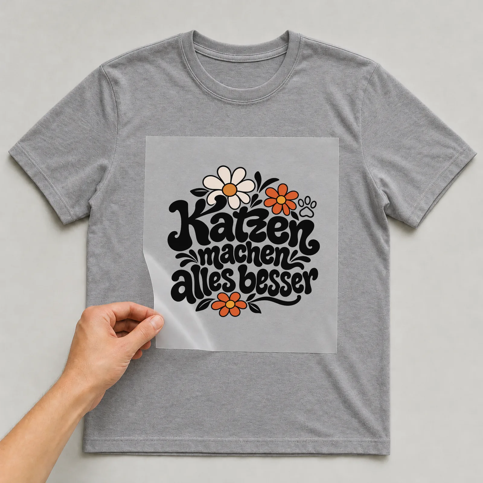 Bügelbild „Katzen machen alles besser“ (Blumenlettering) auf grauem T-Shirt, Folie wird aufgelegt