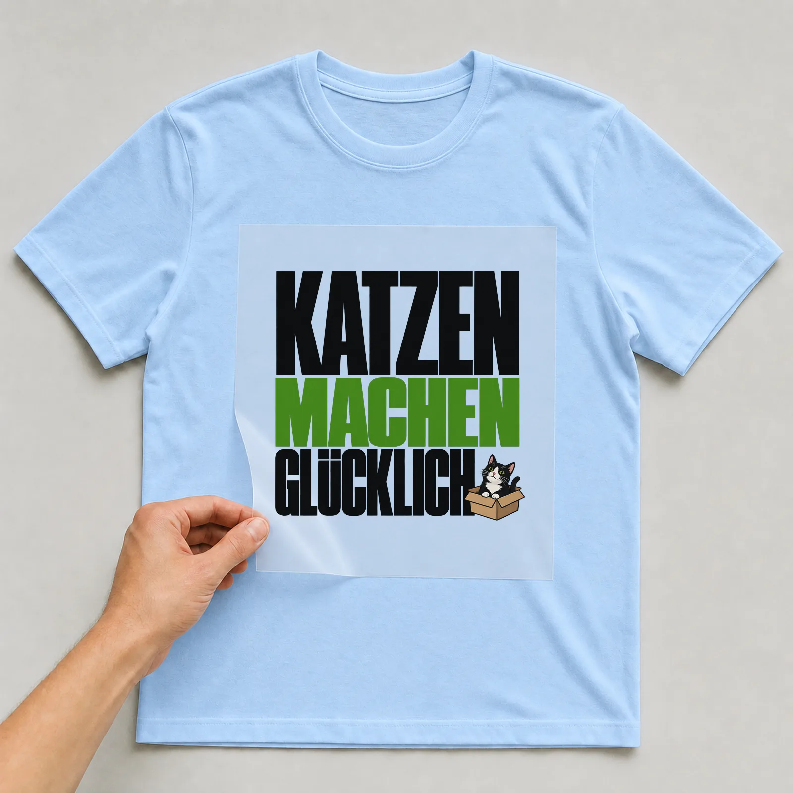 Bügelbild „Katzen machen glücklich“ auf hellblauem T-Shirt, Folie wird aufgelegt