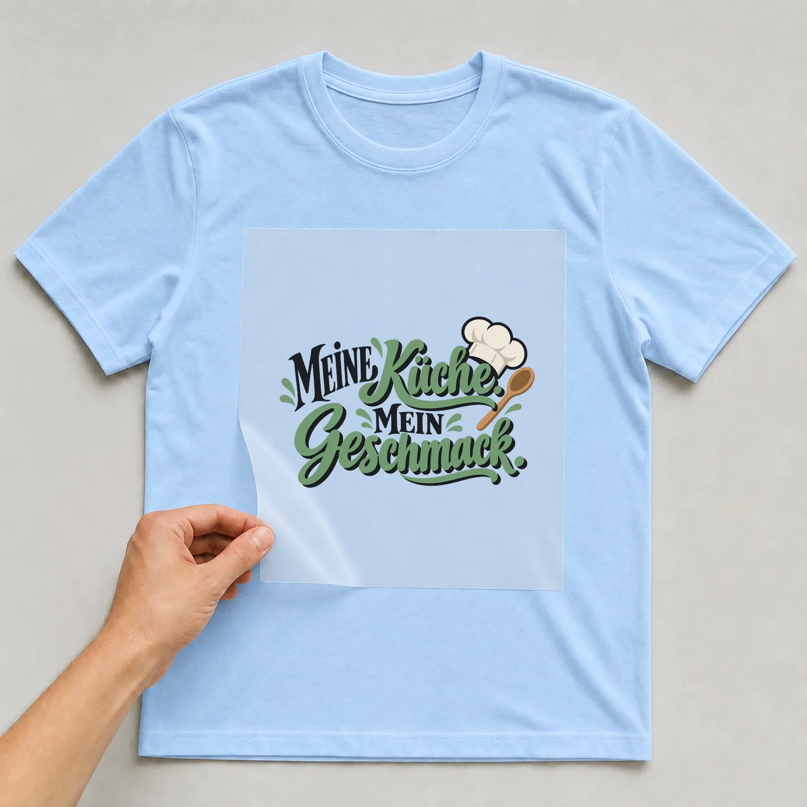 Bügelbild „Meine Küche. Mein Geschmack.“ auf hellblauem T-Shirt, Folie wird aufgelegt