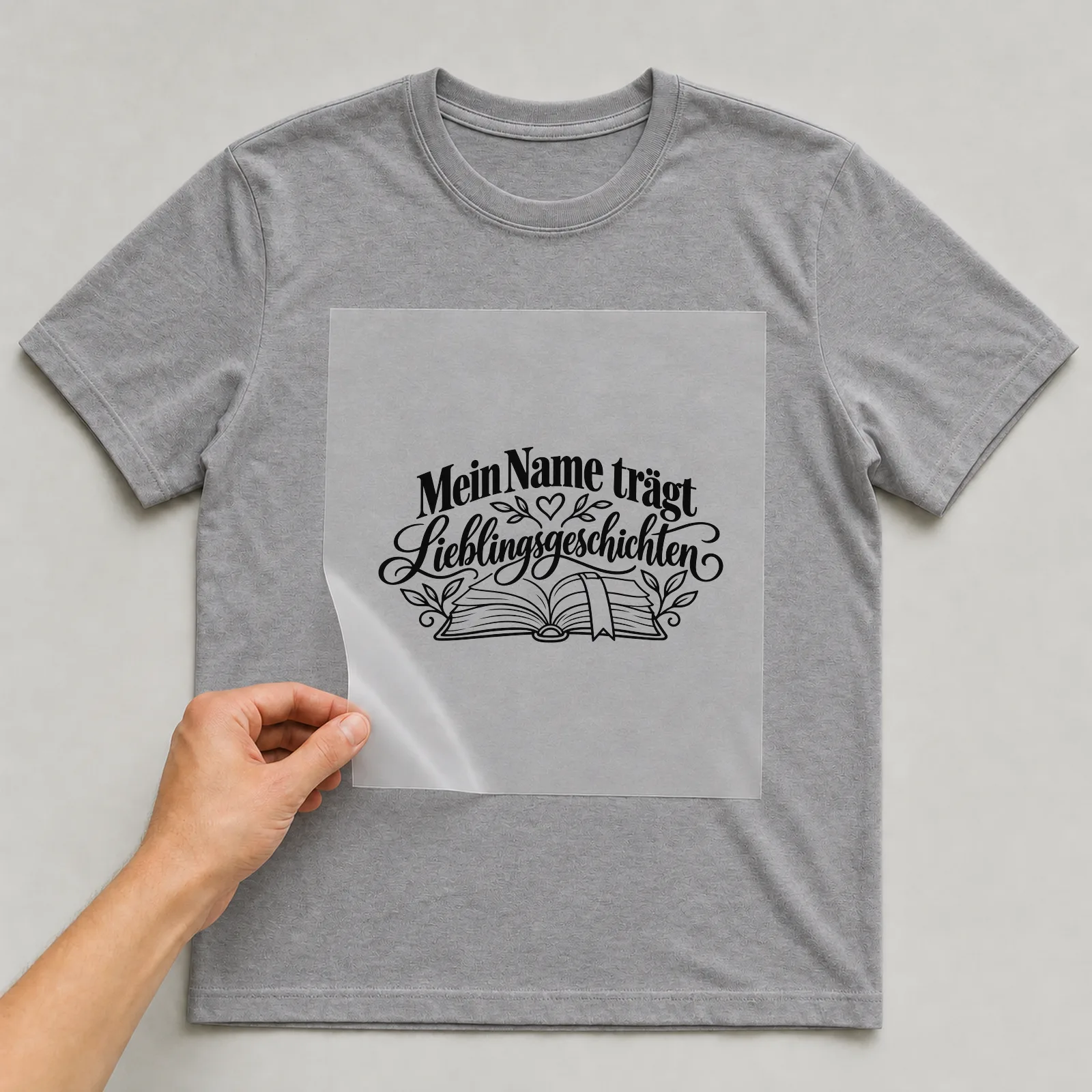 Bügelbild „Mein Name trägt Lieblingsgeschichten“ auf grauem T-Shirt, Folie wird aufgelegt