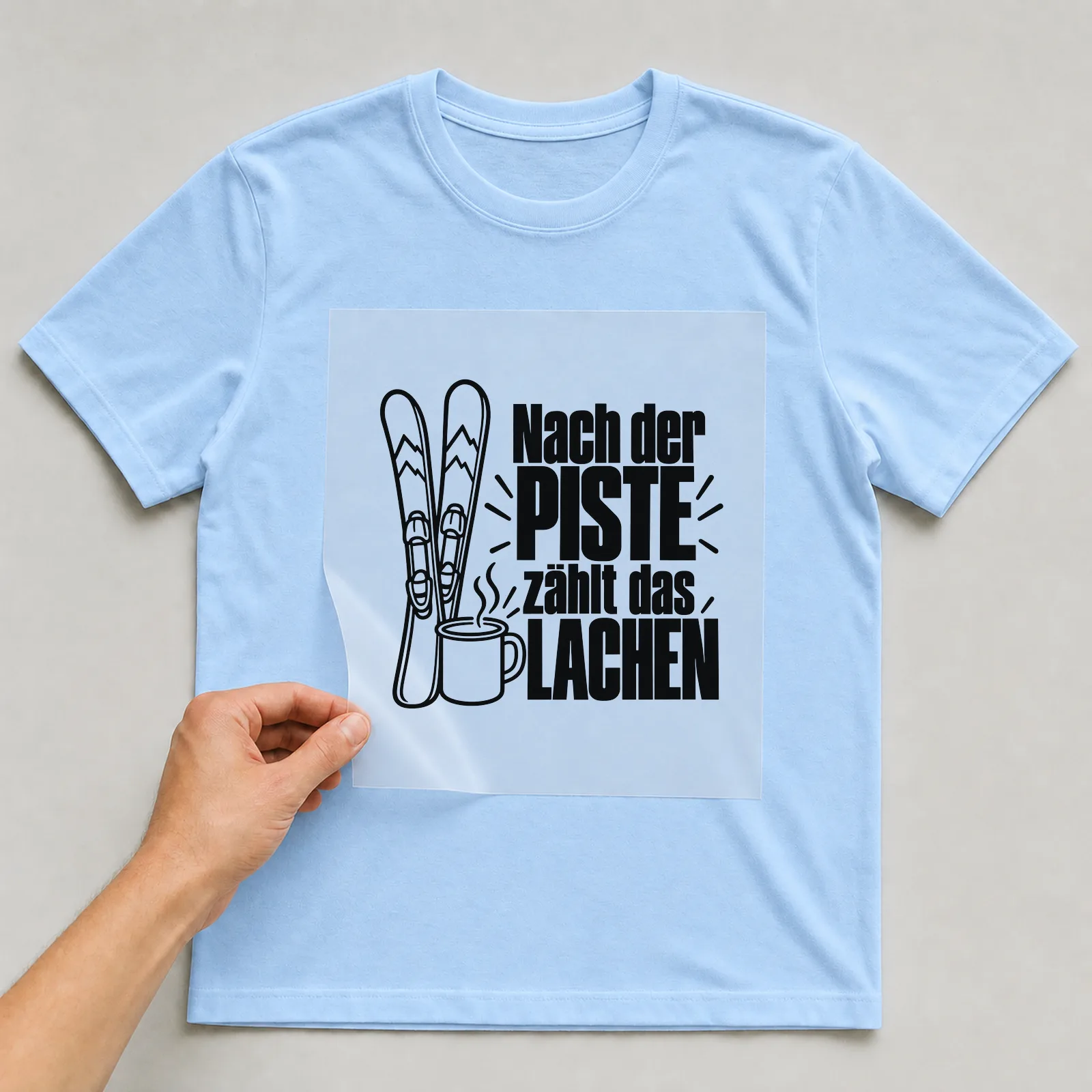 Bügelbild „Nach der Piste zählt das Lachen“ auf hellblauem T-Shirt, Folie wird aufgelegt