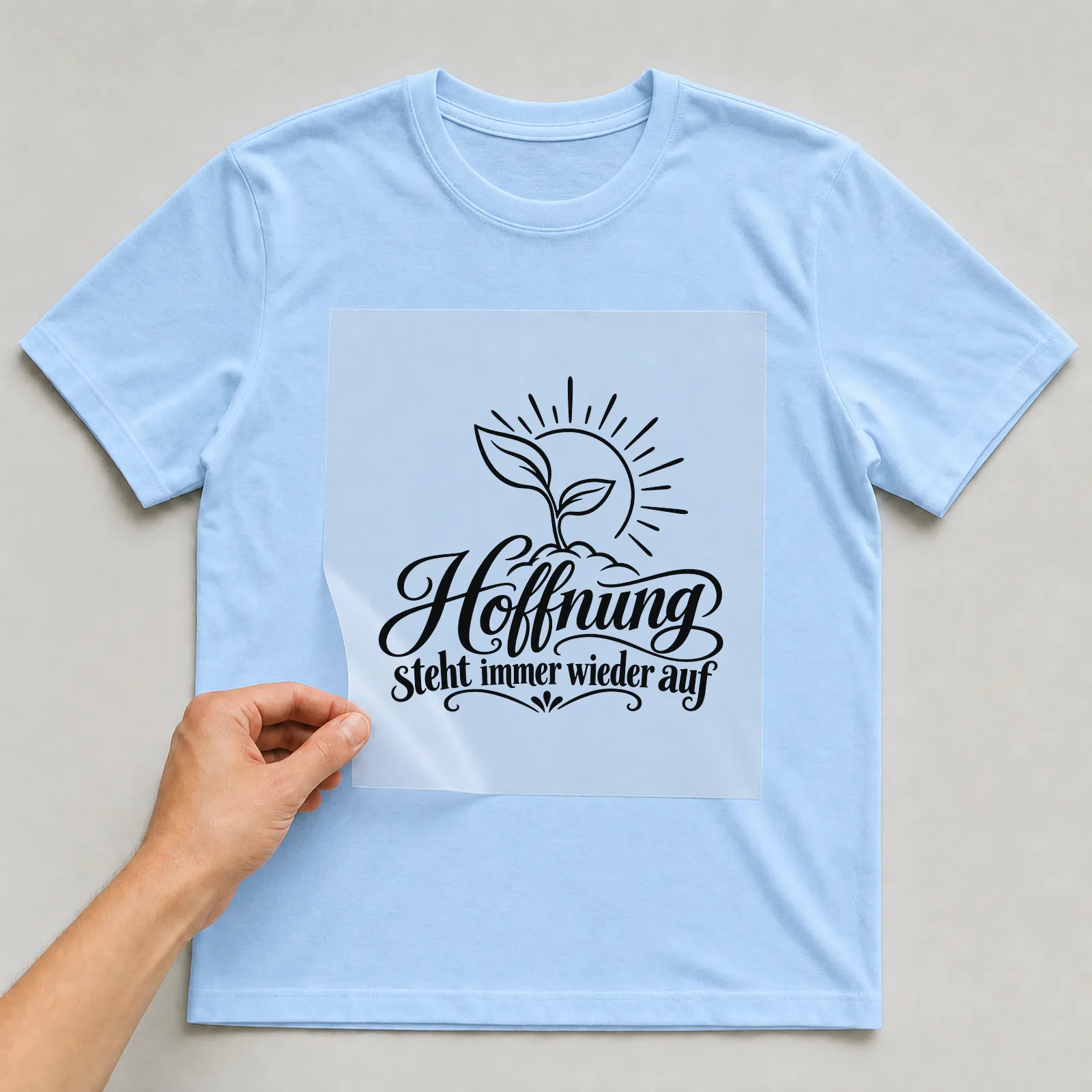Bügelbild „Hoffnung steht immer wieder auf“ auf hellblauem T-Shirt, Folie wird aufgelegt