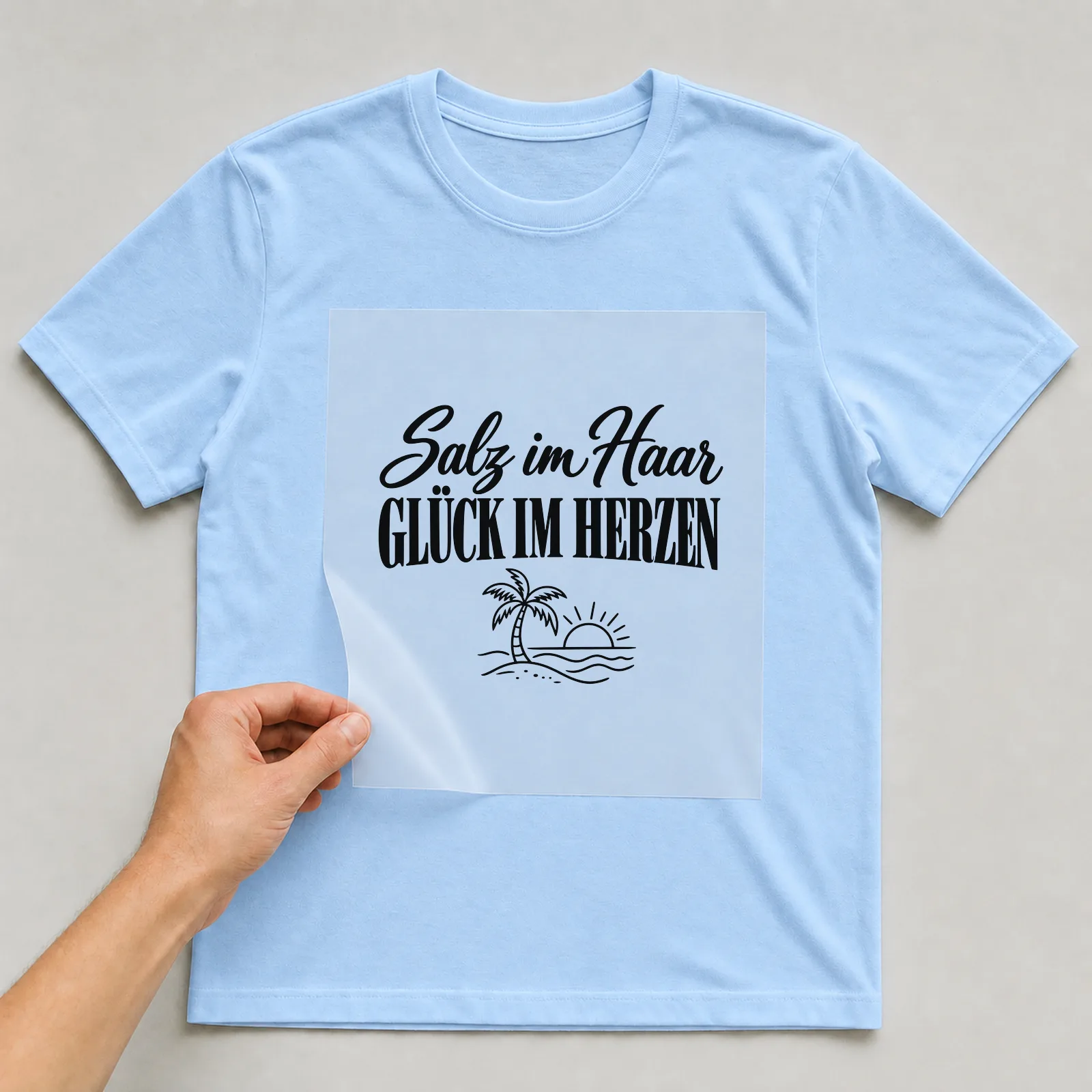 Bügelbild „Salz im Haar Glück im Herzen“ auf hellblauem T-Shirt, Folie wird aufgelegt