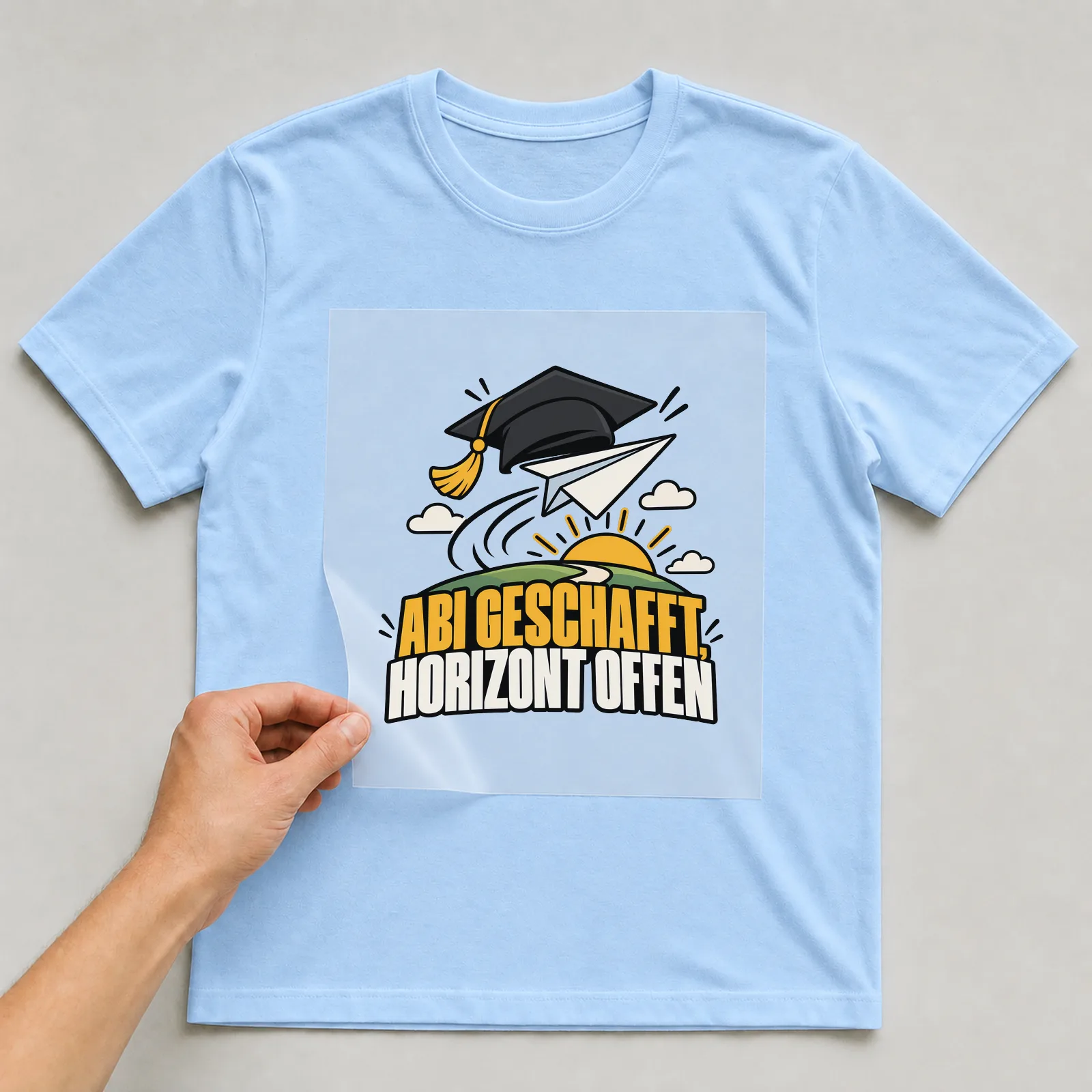 Bügelbild „Abi geschafft, Horizont offen“ auf hellblauem T-Shirt, Folie wird aufgelegt