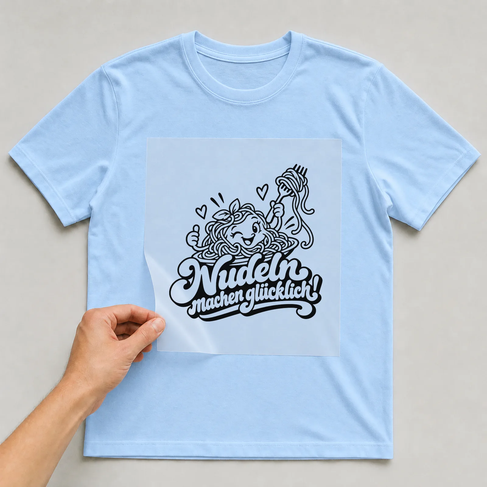 Bügelbild „Nudeln machen glücklich!“ auf hellblauem T-Shirt, Folie wird aufgelegt