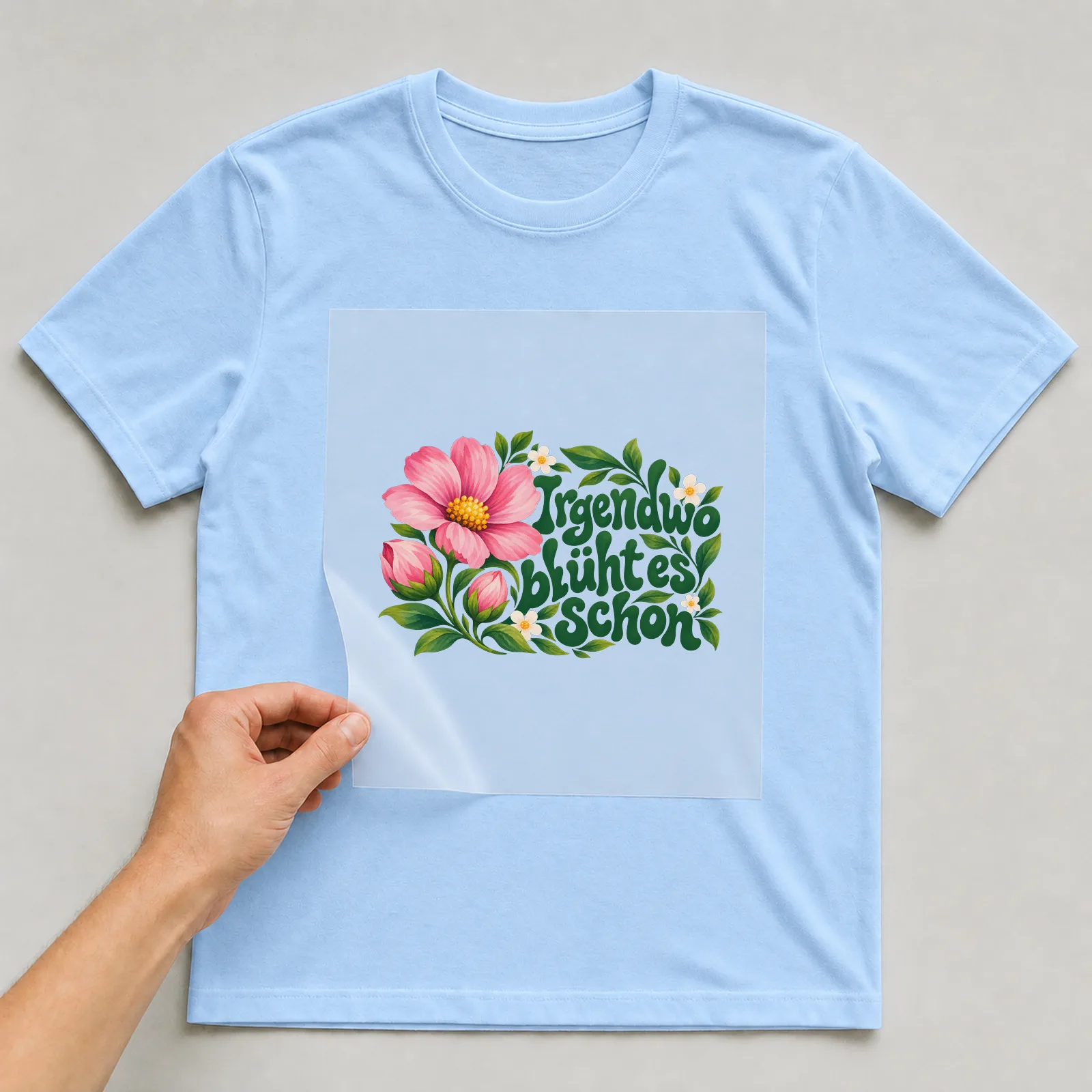 Bügelbild „Irgendwo blüht es schon“ auf hellblauem T-Shirt, Folie wird aufgelegt