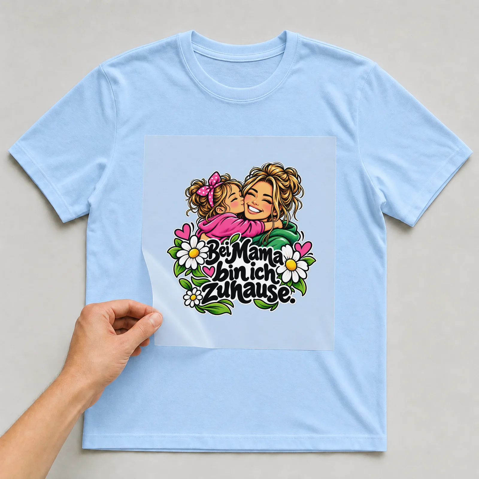 Bügelbild „Bei Mama bin ich zuhause“ auf hellblauem T-Shirt, Folie wird aufgelegt