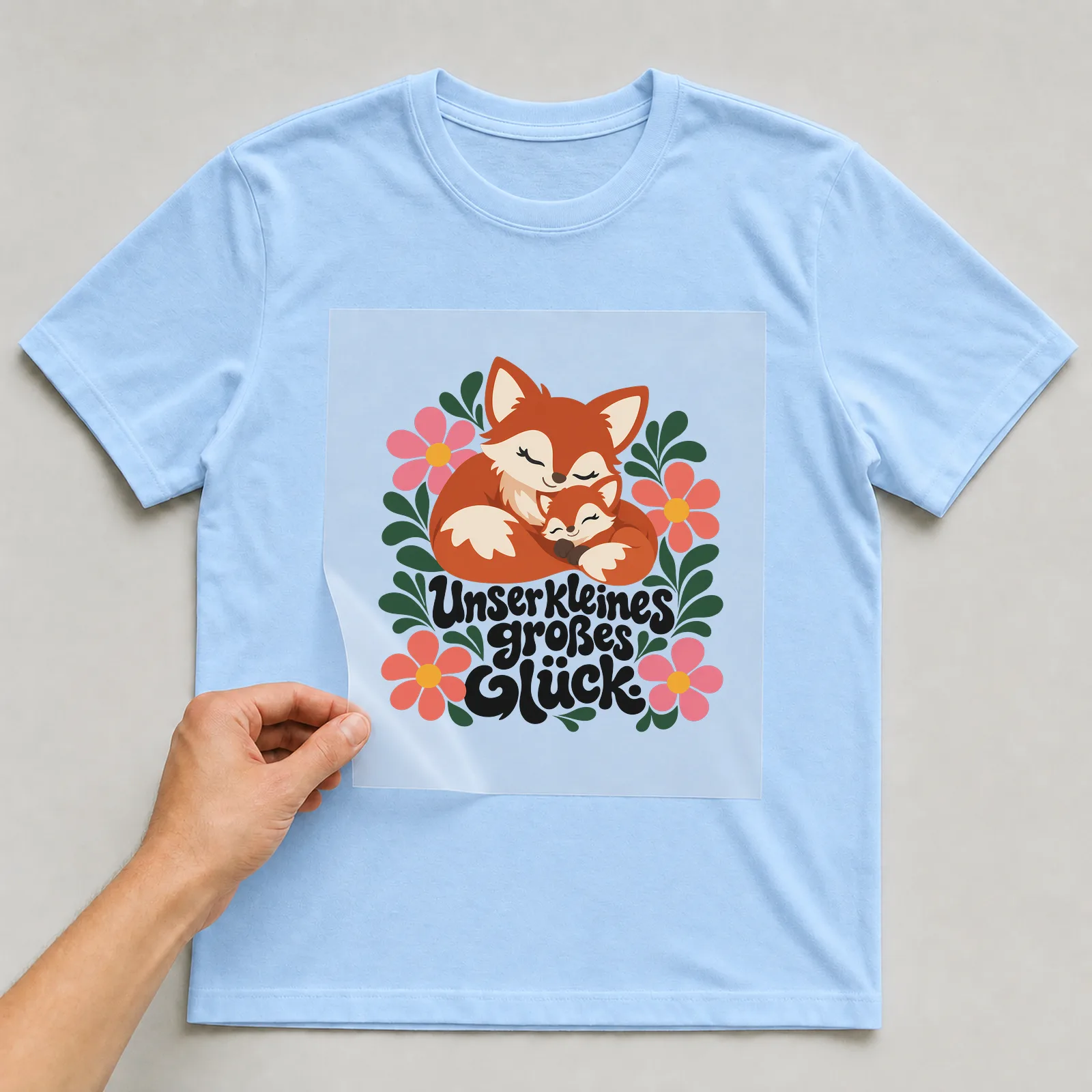 Bügelbild „Unser kleines großes Glück“ auf hellblauem T-Shirt, Folie wird aufgelegt