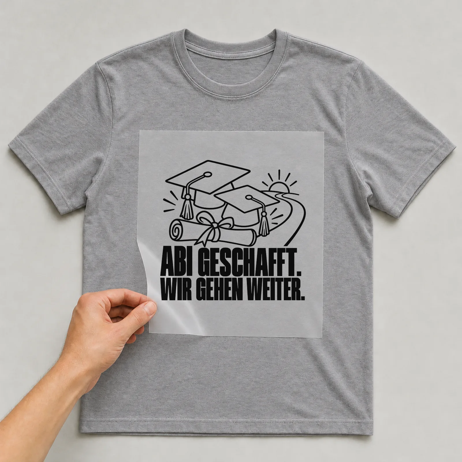 Bügelbild „Abi geschafft. Wir gehen weiter.“ auf grauem T-Shirt, Folie wird aufgelegt