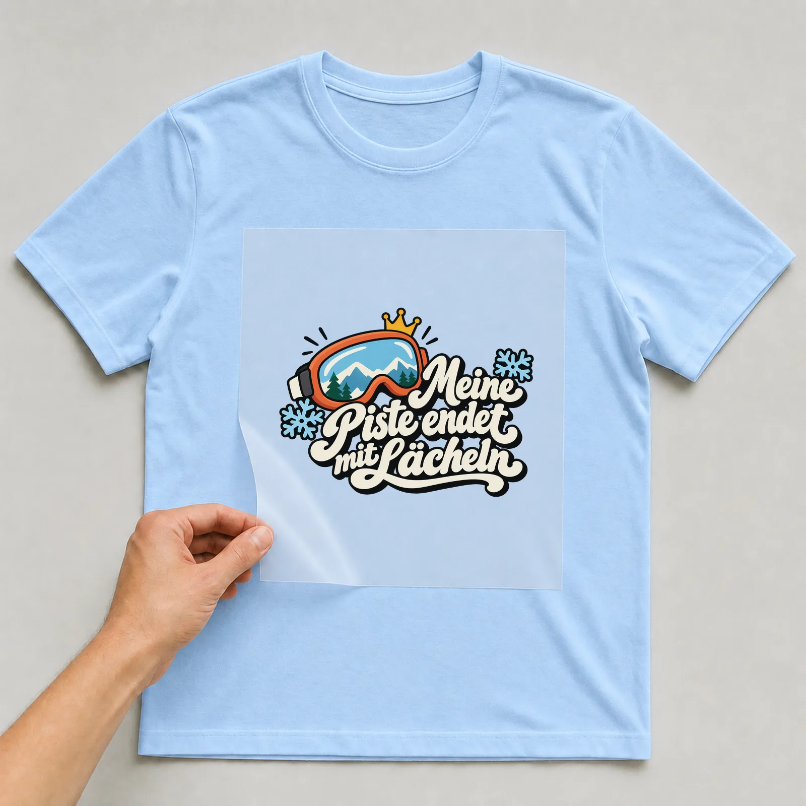 Bügelbild „Meine Piste endet mit Lächeln“ auf hellblauem T-Shirt, Folie wird aufgelegt