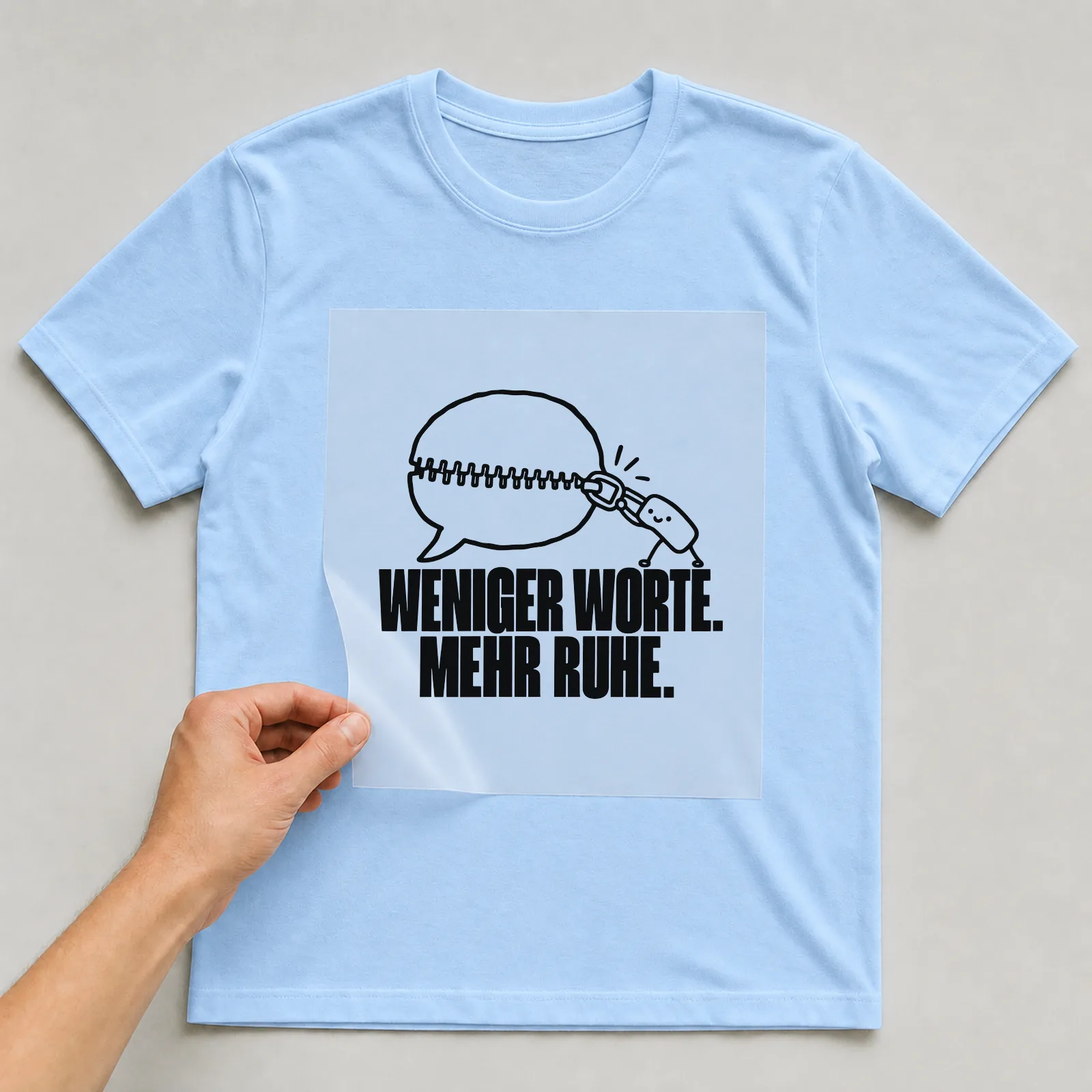 Bügelbild „Weniger Worte. Mehr Ruhe.“ auf hellblauem T-Shirt, Folie wird aufgelegt