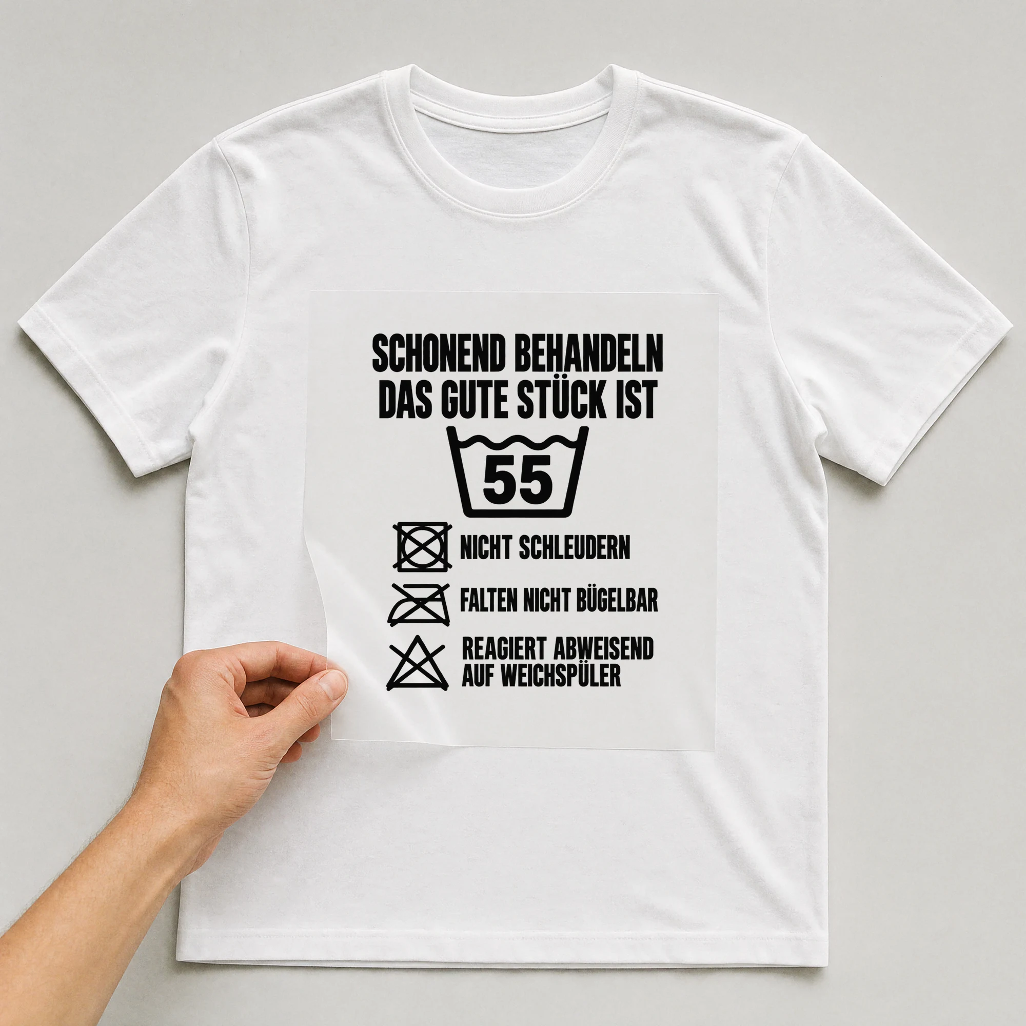 Bügelbild „Schonend behandeln das gute Stück ist 55 nicht schleudern falten nicht“ auf weißem T-Shirt, Folie wird aufgelegt