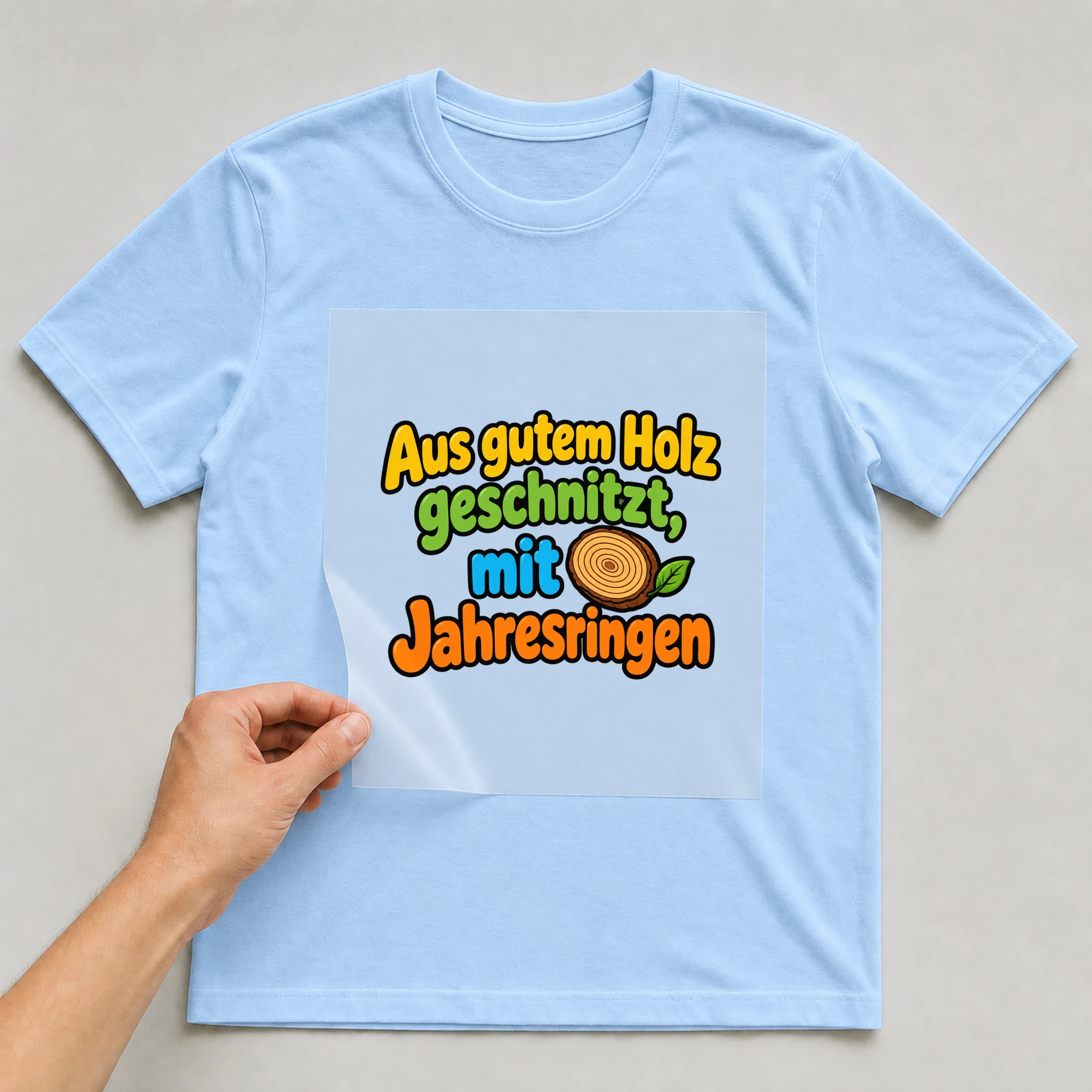 Bügelbild „Aus gutem holz geschnitzt mit jahresringen“ auf babyblauem T-Shirt, Folie wird aufgelegt