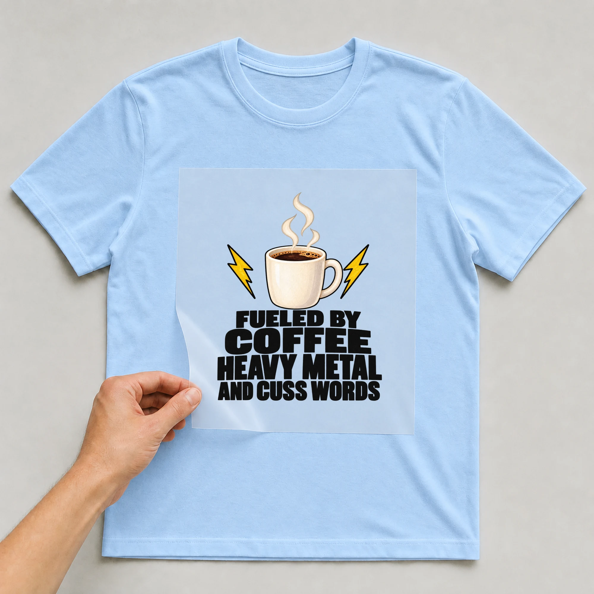 Bügelbild „Fueled By Coffee Heavy Metal And Cuss Words“ auf babyblauem T-Shirt, Folie wird aufgelegt
