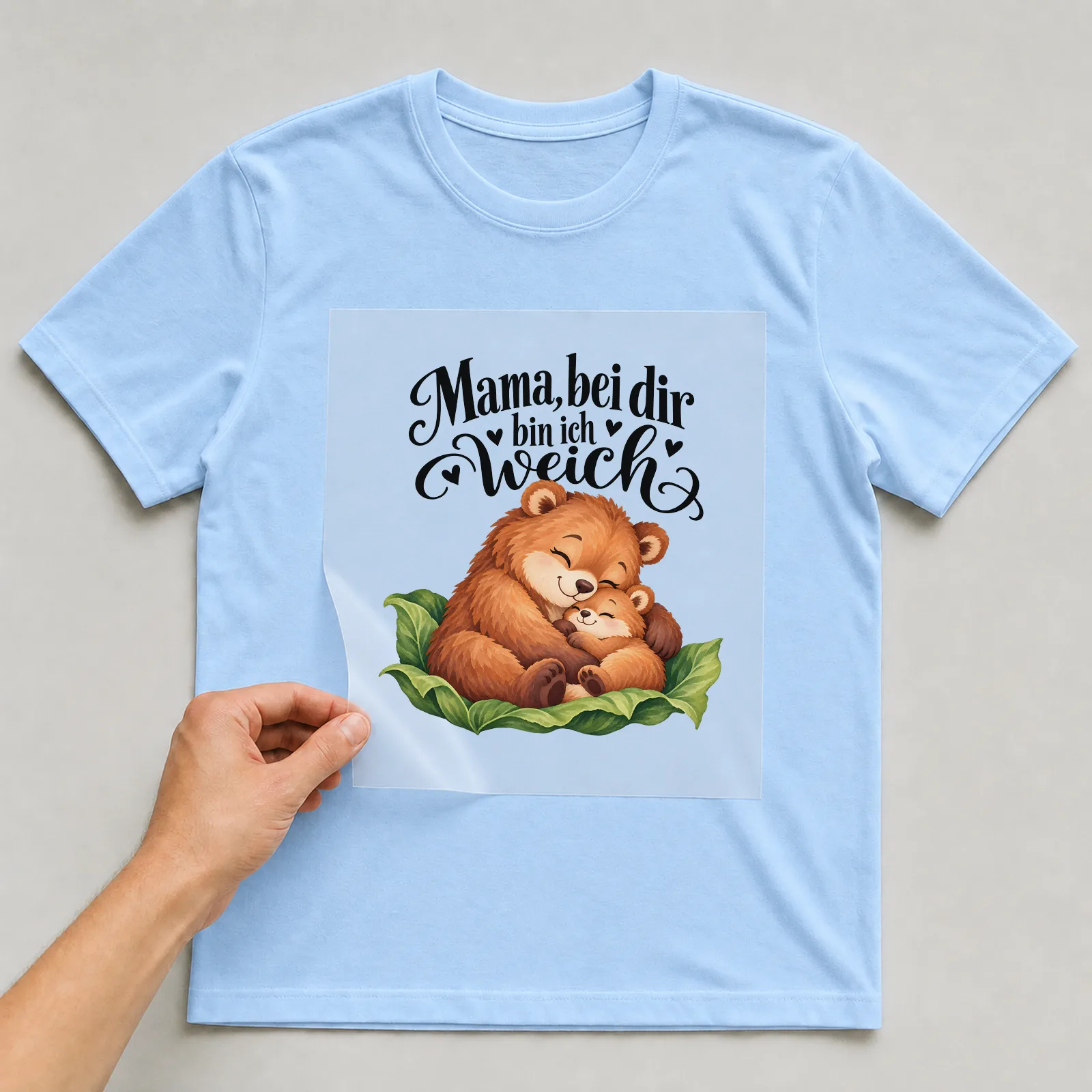 Bügelbild „Mama, bei dir bin ich weich“ auf hellblauem T-Shirt, Folie wird aufgelegt