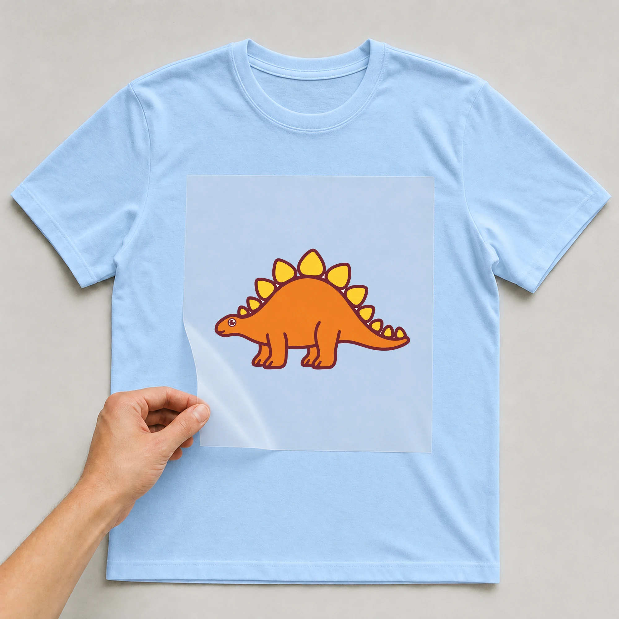 Bügelbild „Dino Stegosaurus“ auf babyblauem T-Shirt, Folie wird aufgelegt