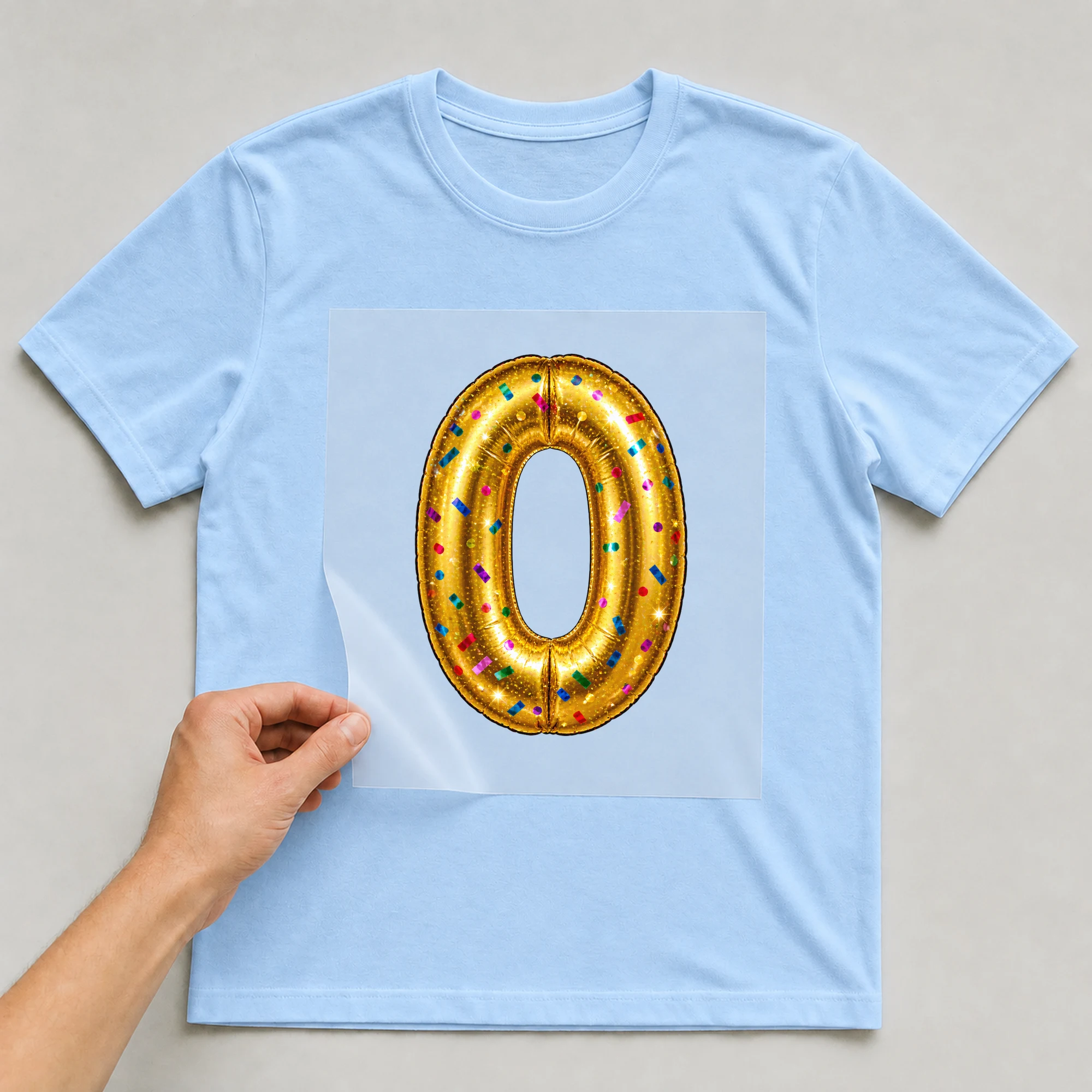 Bügelbild „Zahl 0 im Konfetti-Metallic-Look“ auf babyblauem T-Shirt, Folie wird aufgelegt