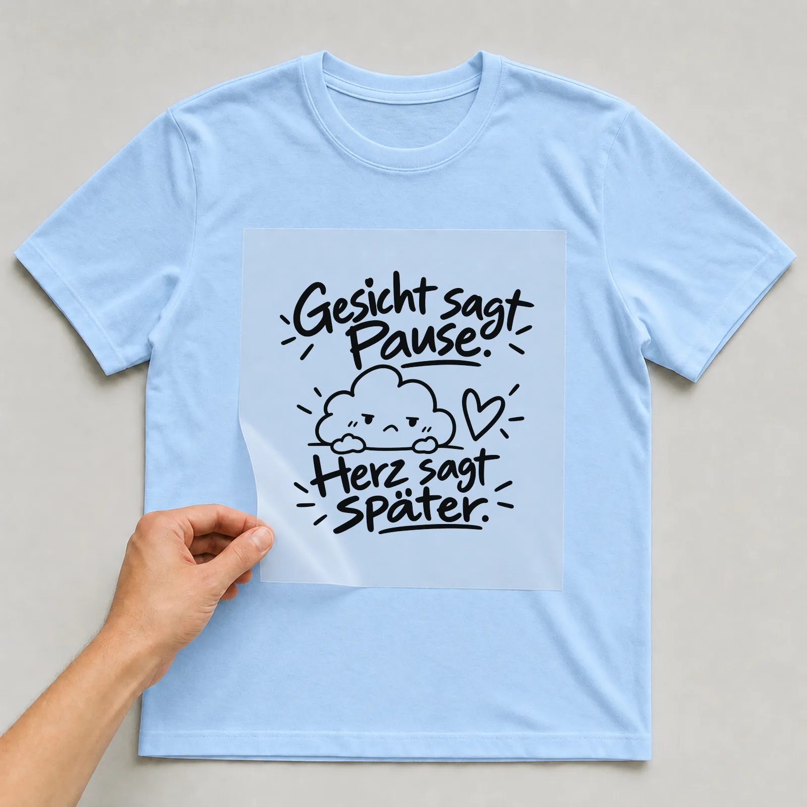 Bügelbild „Gesicht sagt Pause. Herz sagt später.“ auf hellblauem T-Shirt, Folie wird aufgelegt