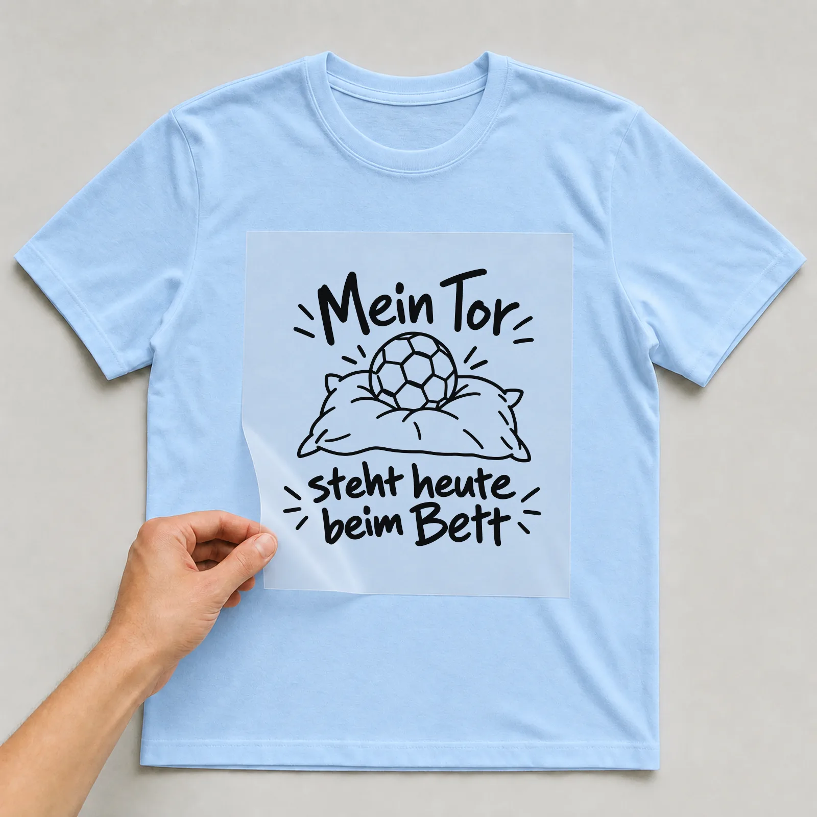 Bügelbild „Mein Tor steht heute beim Bett“ auf hellblauem T-Shirt, Folie wird aufgelegt