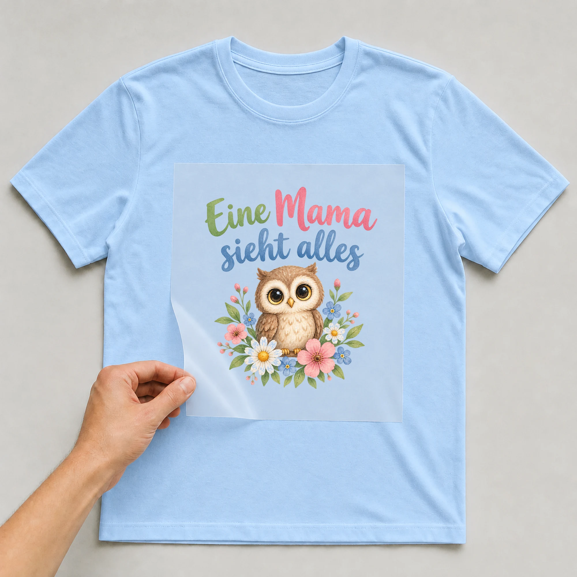 Bügelbild „Eine Mama sieht alles“ auf babyblauem T-Shirt, Folie wird aufgelegt