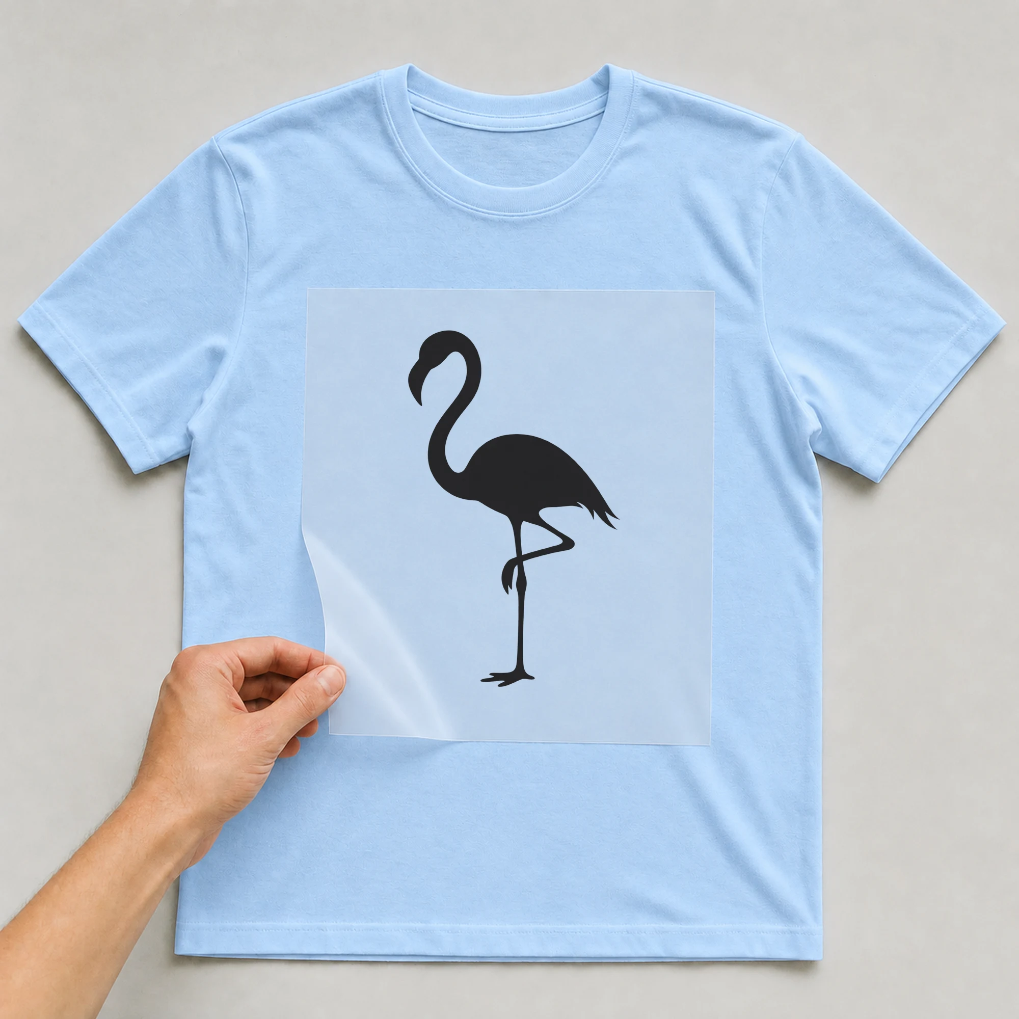 Bügelbild „Flamingo Silhouette“ auf babyblauem T-Shirt, Folie wird aufgelegt