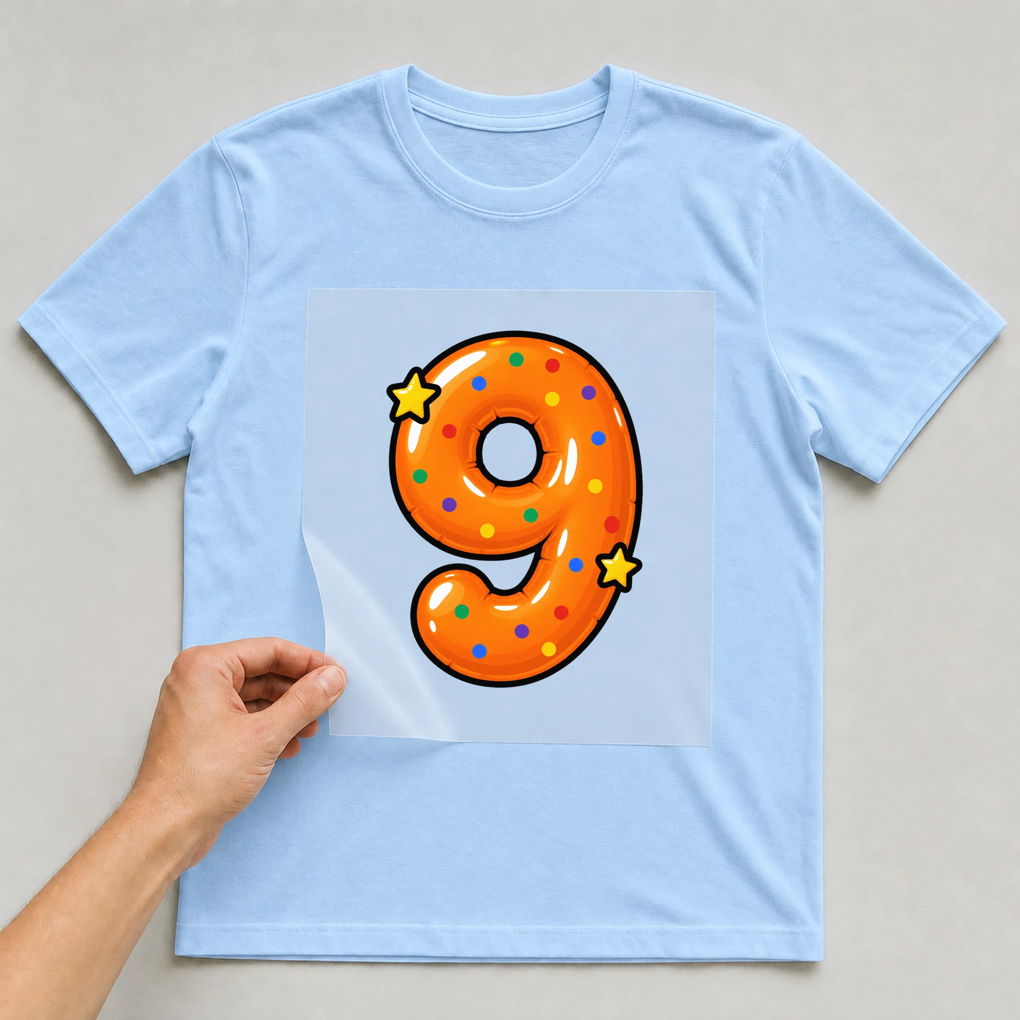 Bügelbild „Zahl 9 im Comic-Ballon-Stil“ auf babyblauem T-Shirt, Folie wird aufgelegt