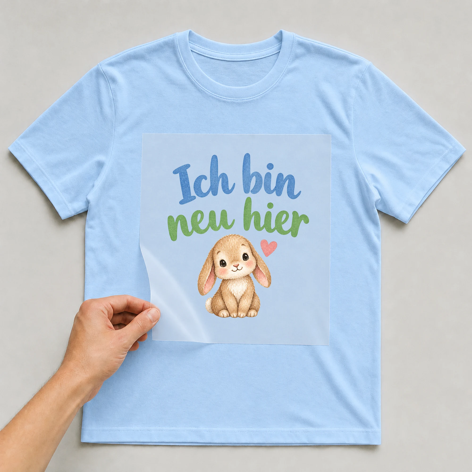 Bügelbild „Ich bin neu hier“ auf babyblauem T-Shirt, Folie wird aufgelegt