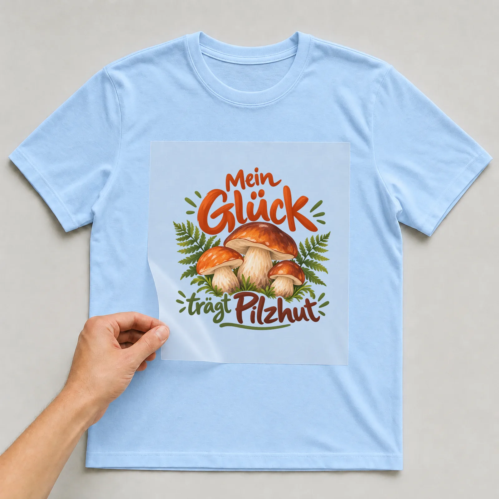 Bügelbild „Mein Glück trägt Pilzhut“ auf hellblauem T-Shirt, Folie wird aufgelegt