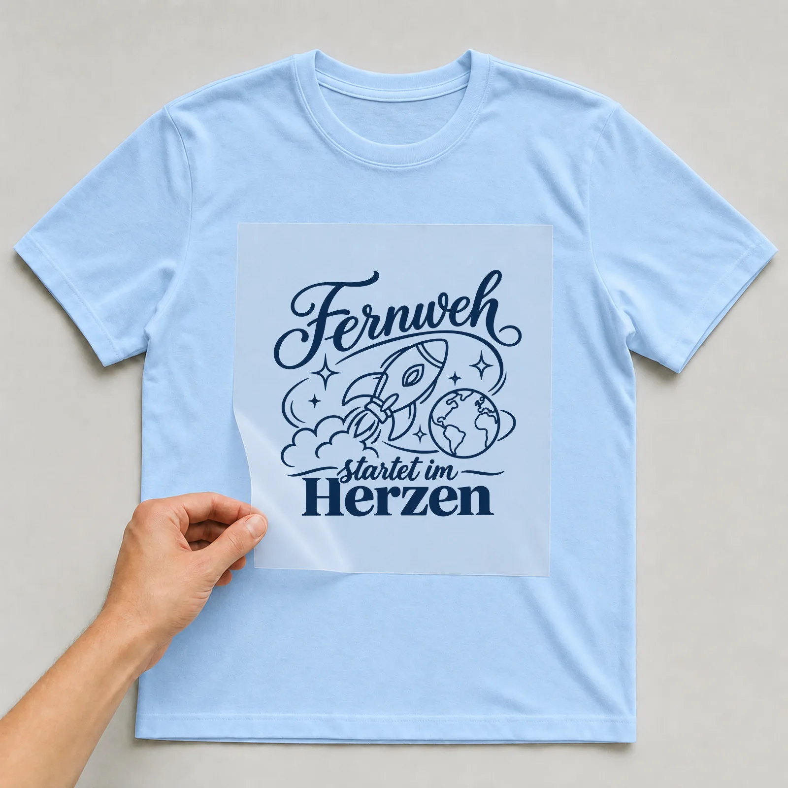 Bügelbild „Fernweh startet im Herzen“ auf hellblauem T-Shirt, Folie wird aufgelegt