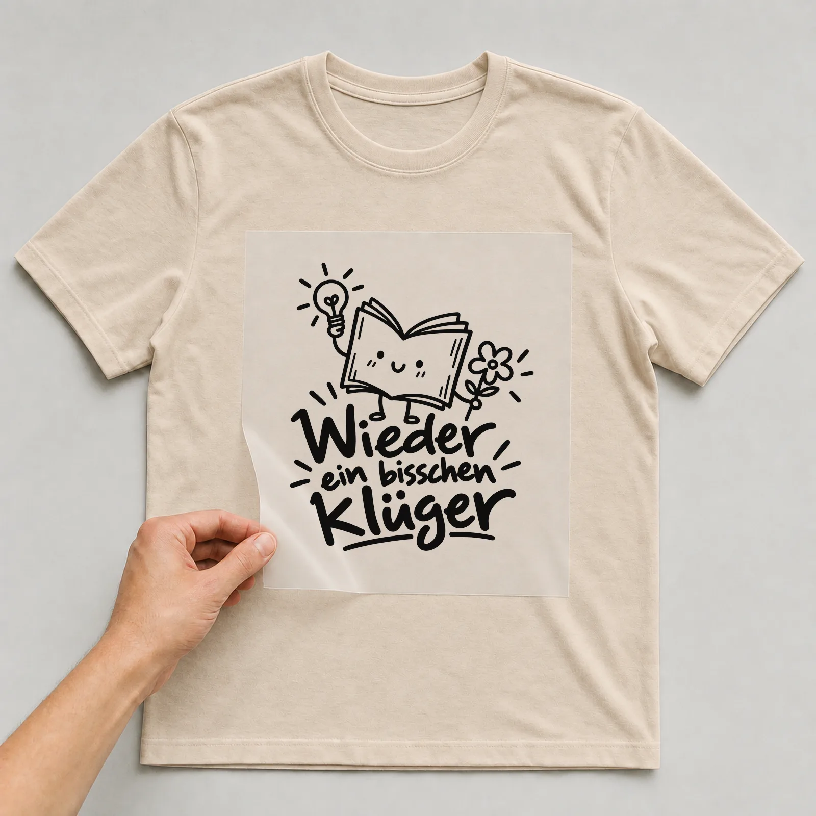 Bügelbild „Wieder ein bisschen klüger“ auf beigem T-Shirt, Folie wird aufgelegt