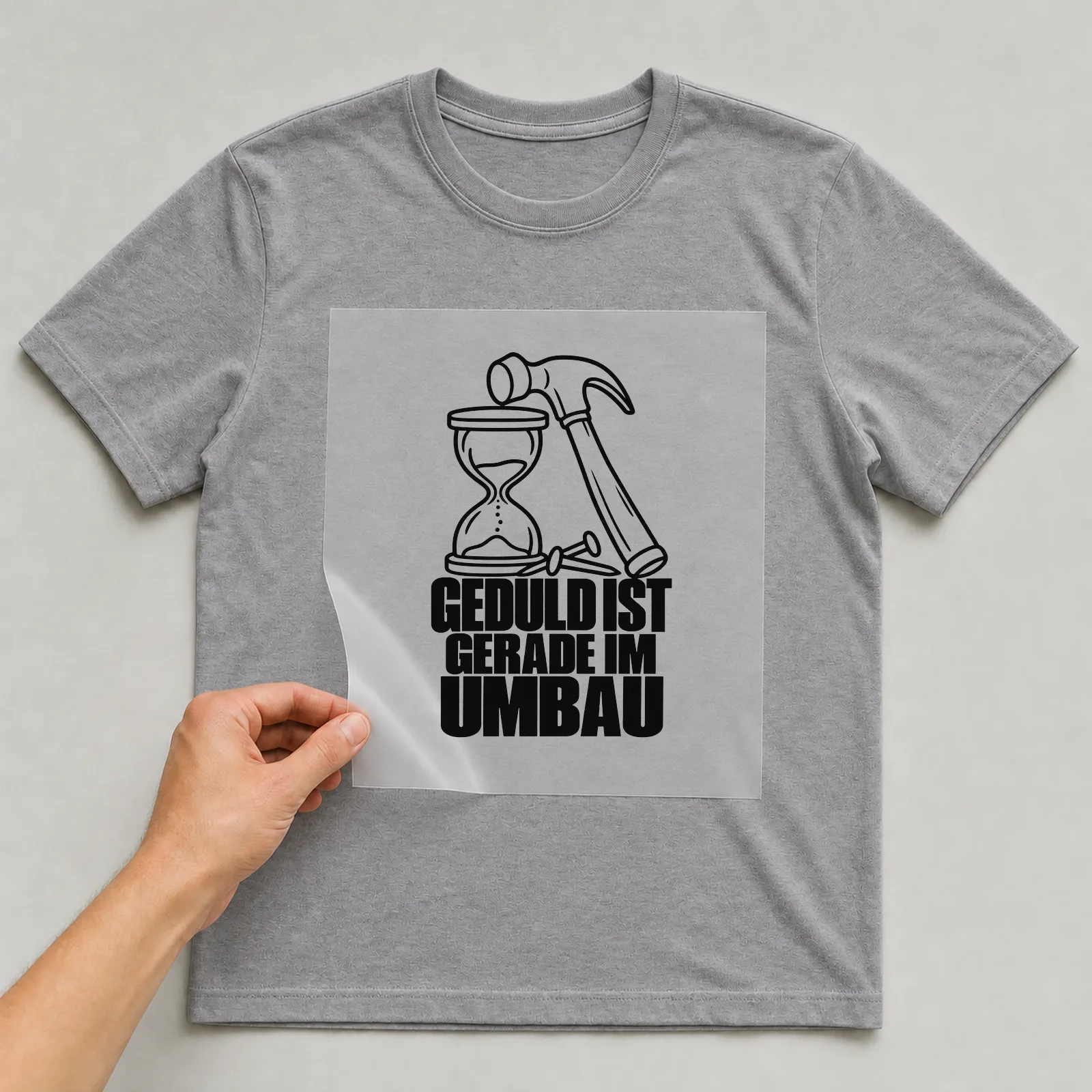 Bügelbild „Geduld ist gerade im Umbau“ auf grauem T-Shirt, Folie wird aufgelegt