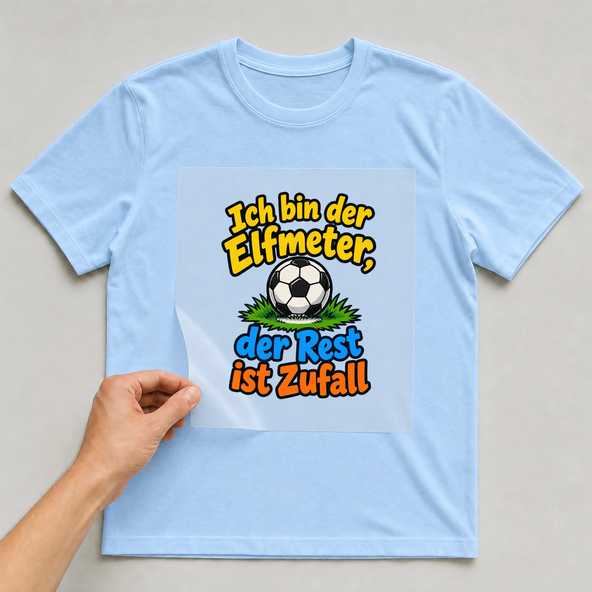 Bügelbild „Ich bin der Elfmeter, der Rest ist Zufall“ auf babyblauem T-Shirt, Folie wird aufgelegt