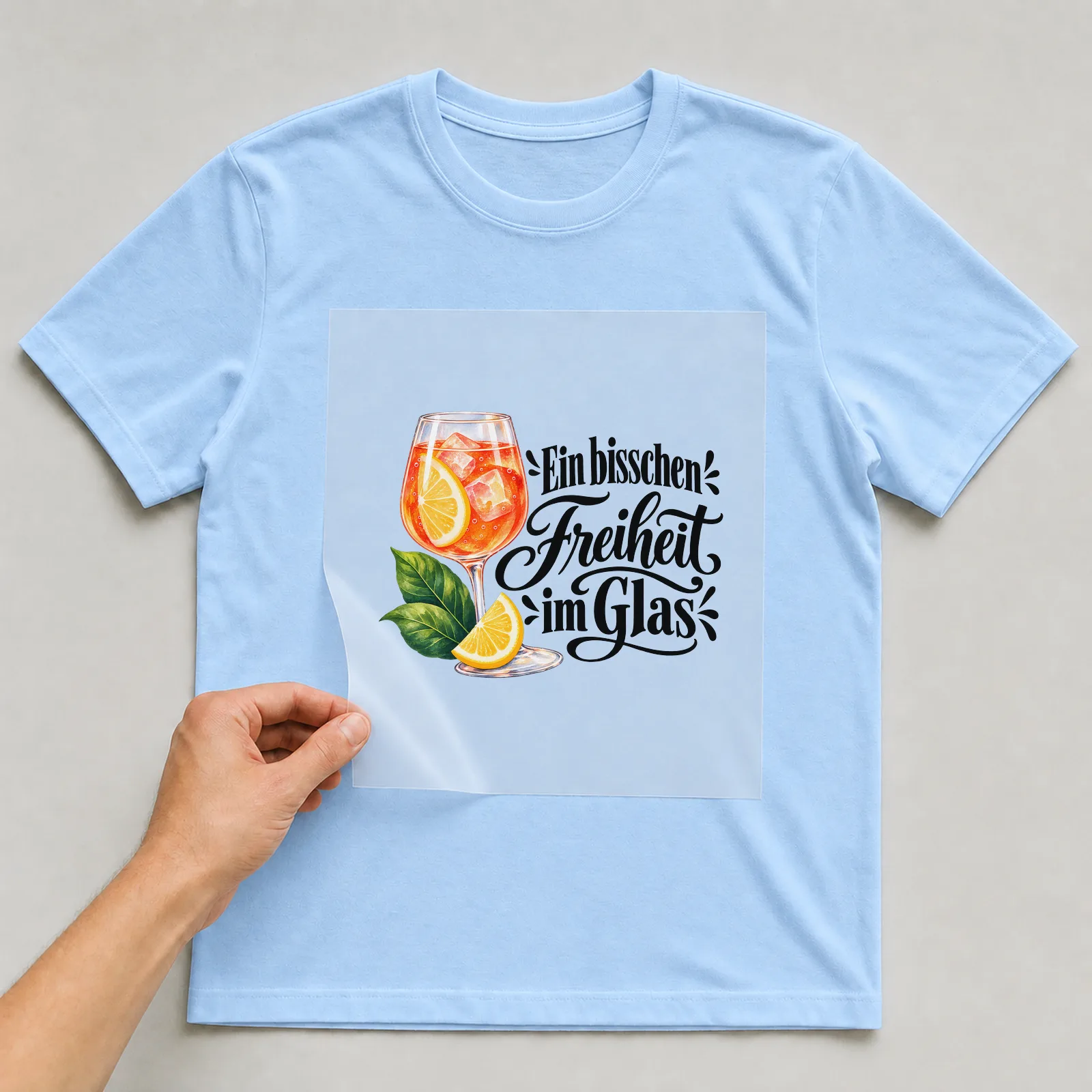 Bügelbild „Ein bisschen Freiheit im Glas“ auf hellblauem T-Shirt, Folie wird aufgelegt